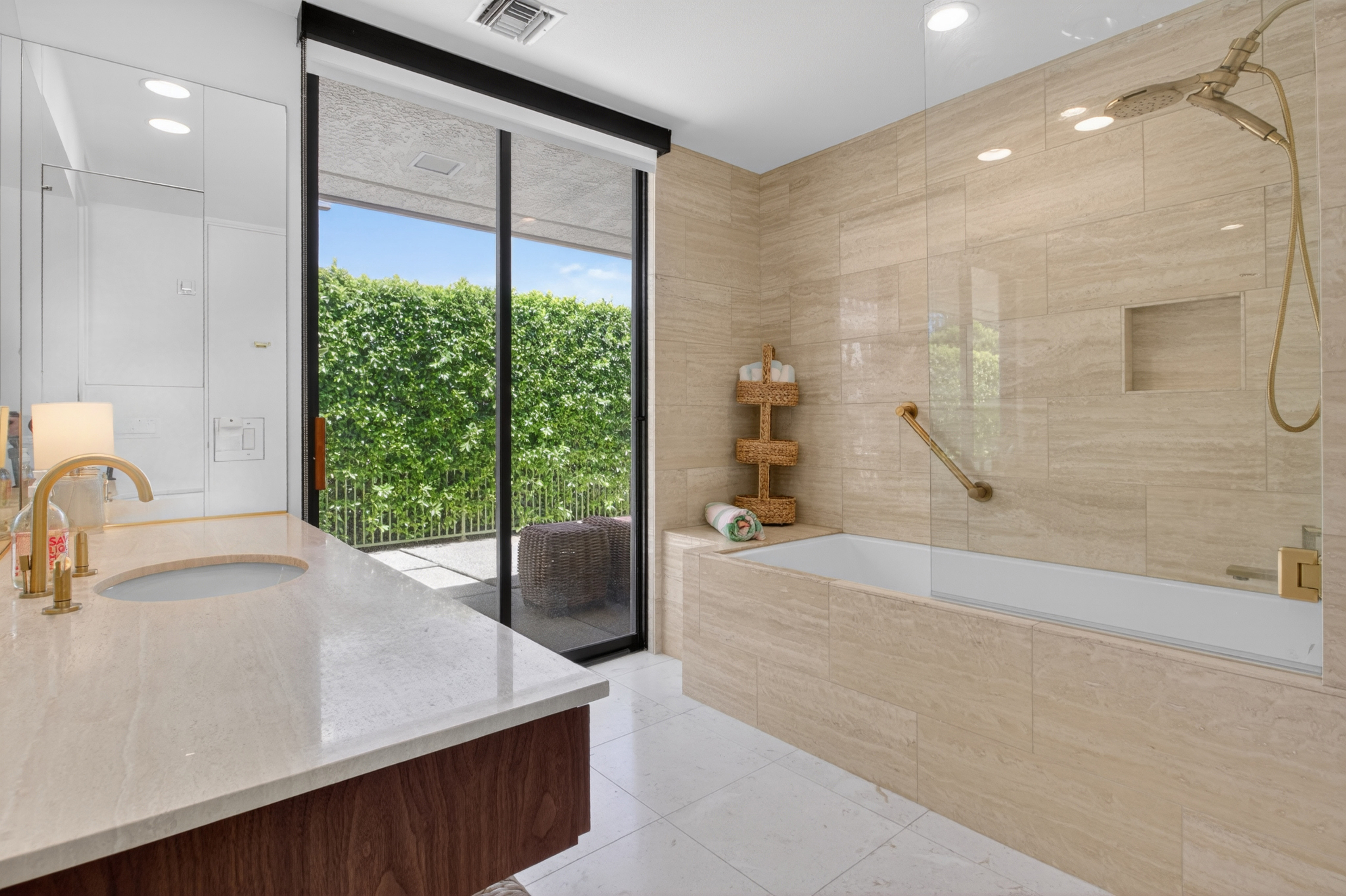This luxurious primary bathroom features a sleek, integrated stone vanity with gold fixtures and a spacious soaking tub combined with a shower. Large-format beige stone tiles create a cohesive, spa-like aesthetic, while floor-to-ceiling glass doors provide a seamless transition to a private, lushly landscaped patio. The room is bright and airy, blending elegant materials with a modern, indoor-outdoor design.