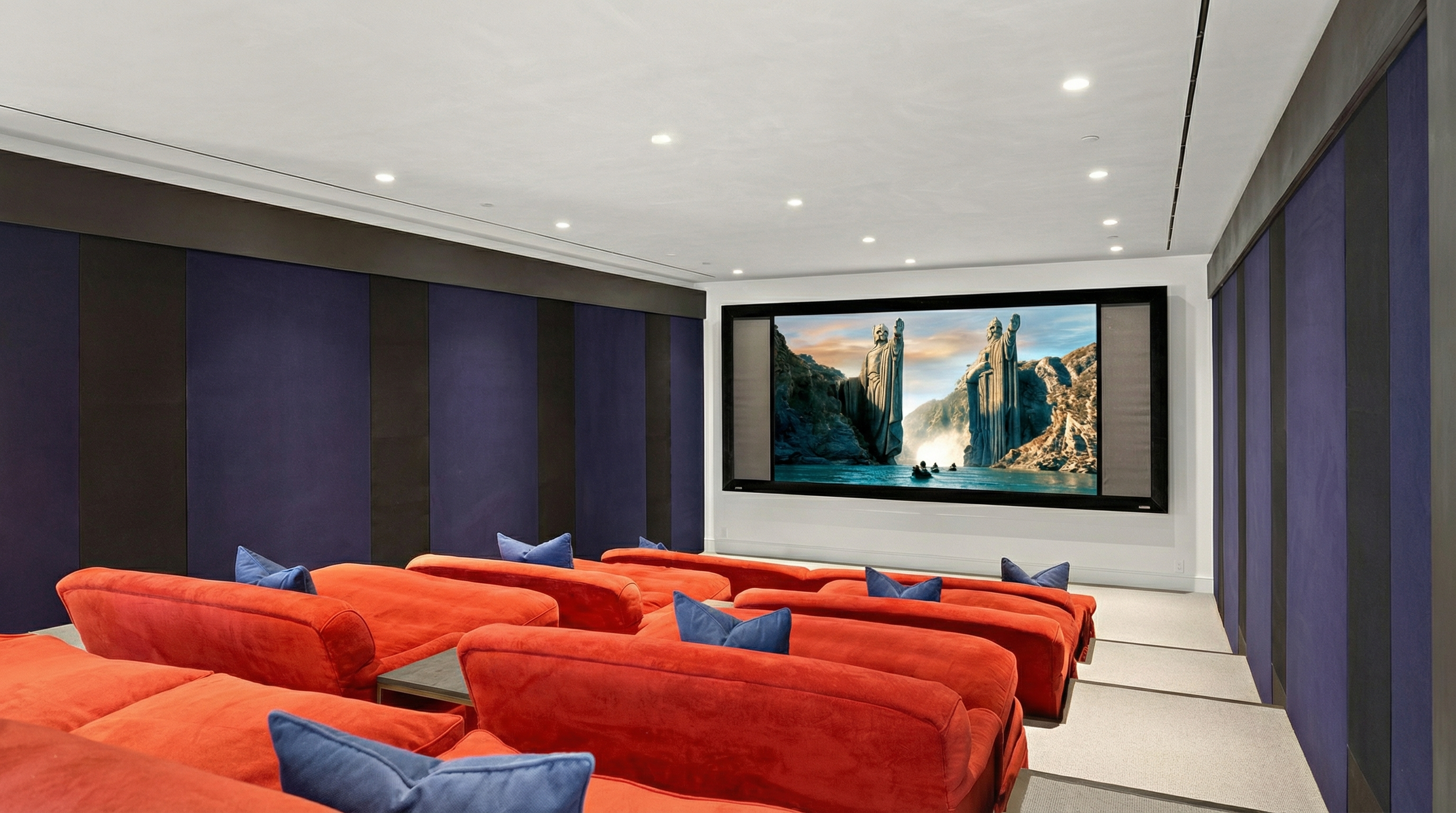 This is an image of a home theater. The room features several rows of plush, red seating, each adorned with blue pillows, facing a large screen displaying a scenic image. Dark blue and gray acoustic panels line the walls, and recessed lighting illuminates the ceiling, creating a comfortable and immersive entertainment space.