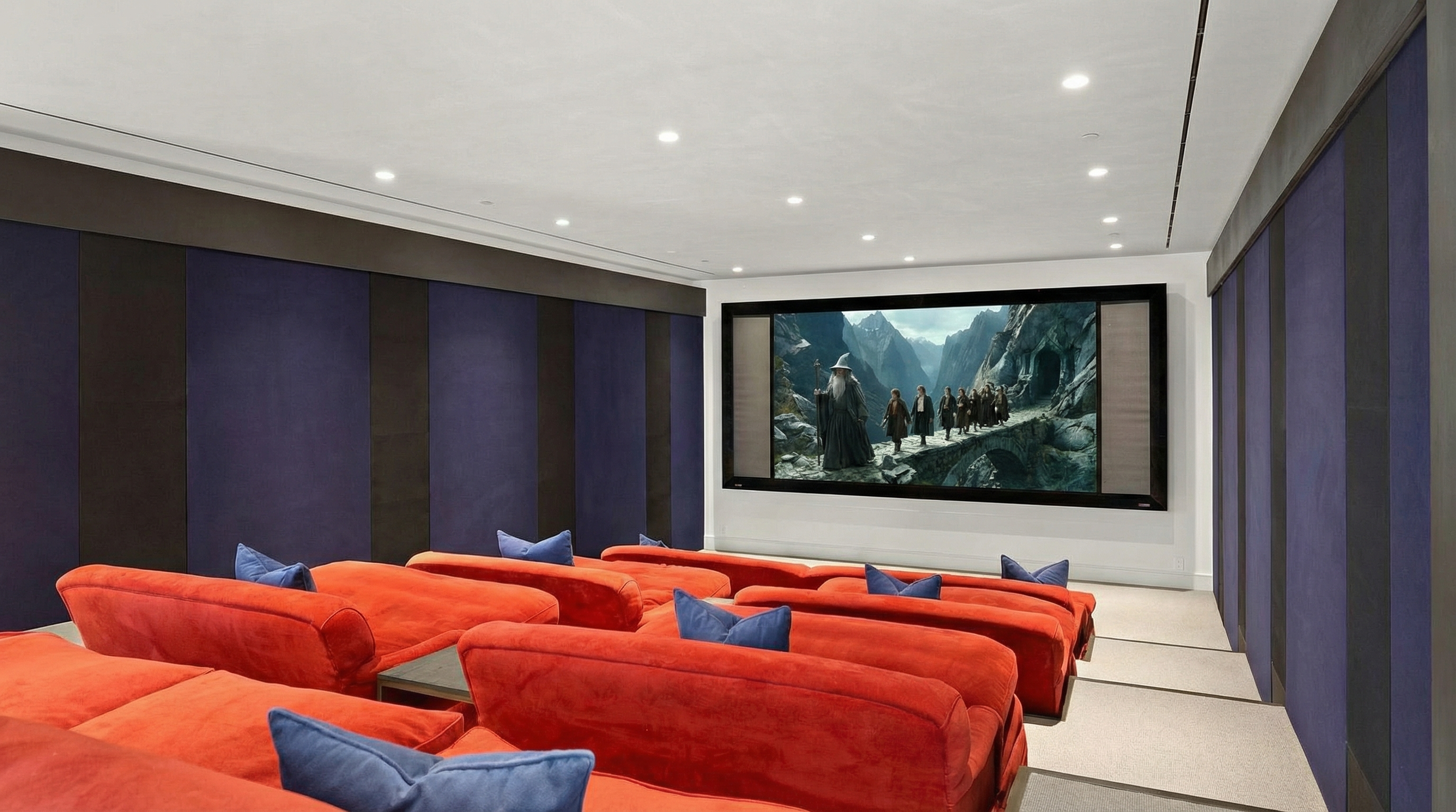 This is a home theater featuring multiple rows of plush red seating with blue throw pillows, perfect for comfortable movie viewing. A large screen displays a scene from "The Lord of the Rings". The walls are paneled with a combination of dark and light tones, contributing to the room's sound insulation and overall aesthetic.
