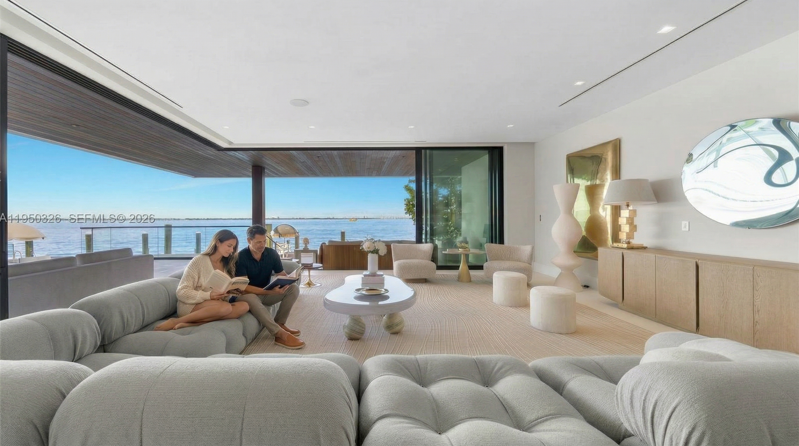 This modern, open-concept living room offers breathtaking, unobstructed waterfront views through floor-to-ceiling glass walls that blend indoor and outdoor living. The space is elegantly furnished with a plush, modular grey sectional, a unique organic-shaped coffee table, and minimalist decor, creating a sophisticated and serene atmosphere. Its expansive layout, neutral color palette, and high-end design elements highlight the property's luxury and emphasis on tranquil, high-end coastal living.