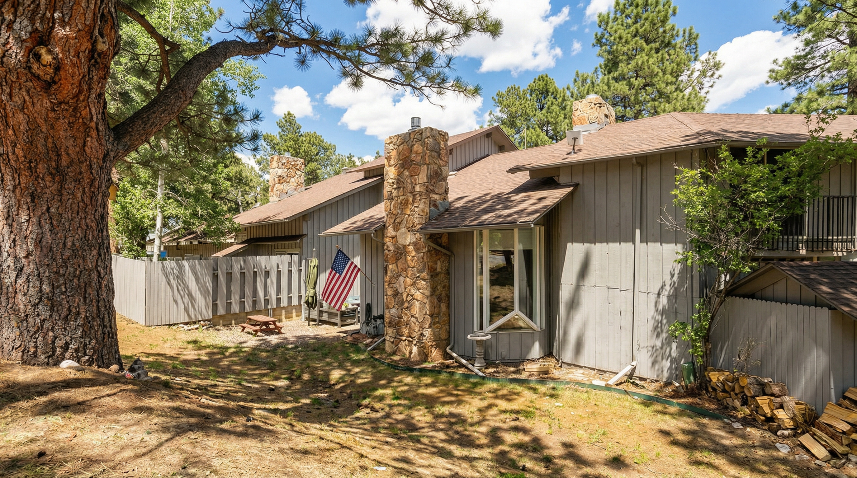 This exterior view showcases the rear of a multi-story home featuring gray vertical siding, a prominent natural stone chimney, and a bay window. The property includes a small, fenced-in patio area adorned with an American flag and a wood pile, nestled among tall pine trees. The rustic, mountain-style aesthetic creates a cozy and serene atmosphere suitable for a woodland setting.