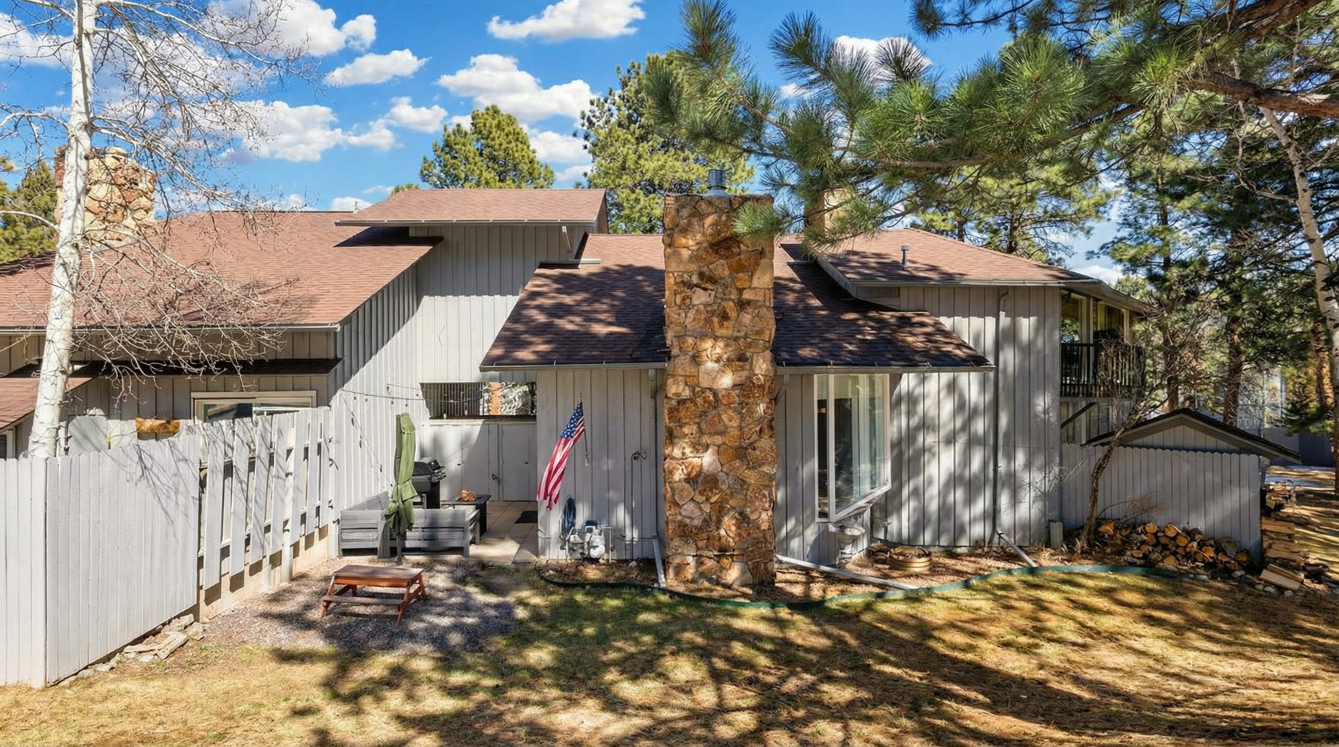This exterior rear view showcases a multi-level house with vertical gray wood siding and a prominent stone chimney. The backyard area includes a small patio space with outdoor seating, an American flag, and a convenient woodshed structure. The surrounding pine trees and natural landscape contribute to a cozy, woodland cabin aesthetic.