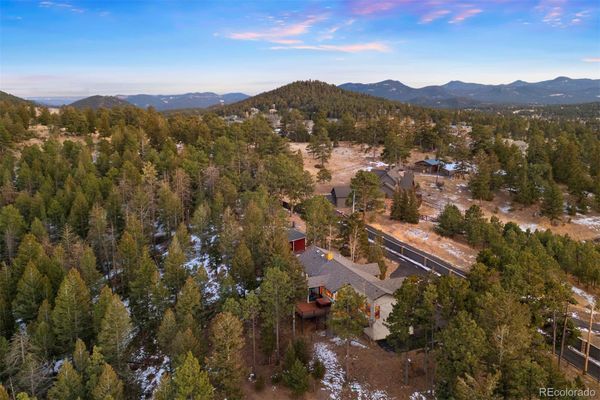 This is an aerial view capturing a residential property nestled within a wooded, mountainous landscape. The scene highlights the home's integration into the forest setting, featuring a spacious lot with a mix of pine trees, patches of snow, and natural terrain. The elevated perspective showcases the surrounding topography and the access road, providing a sense of seclusion and serenity.
