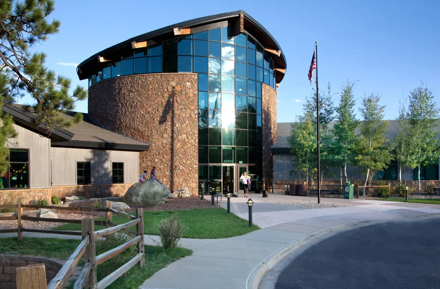 This building features a prominent, circular stone rotunda with a high, glass-curtain wall entrance, presenting a modern and architecturally striking facade. The property includes a paved entryway, neatly landscaped grounds with mature trees, and a rustic wood fence. Its design suggests a professional or public facility, blending natural stone elements with contemporary materials for a welcoming and well-maintained appearance.