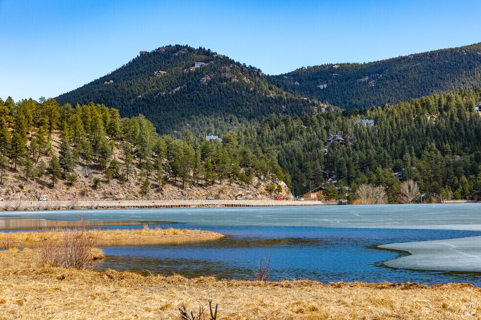 This scenic landscape photograph captures a frozen lake reflecting the surrounding mountainous terrain and dense coniferous forest. The foreground features golden, dormant marsh grasses leading to a partially melted shoreline, while the backdrop showcases distant hillside homes integrated into the natural environment. The image emphasizes the tranquil, natural beauty and outdoor recreational appeal of the location.