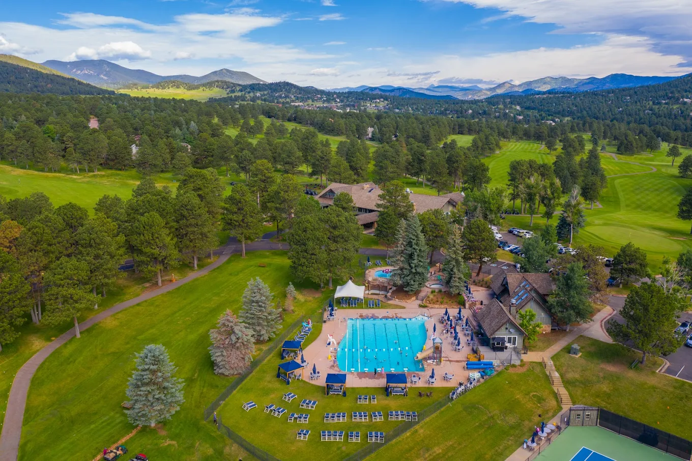 This stunning aerial view captures a premier resort-style property nestled within a lush, mountainous landscape. The expansive grounds feature a large, crystal-clear swimming pool with an adjacent clubhouse and tennis court, all set against a backdrop of rolling green hills and majestic mountain ranges. The property offers an inviting blend of recreational facilities and scenic natural beauty, making it a highly desirable community amenity.