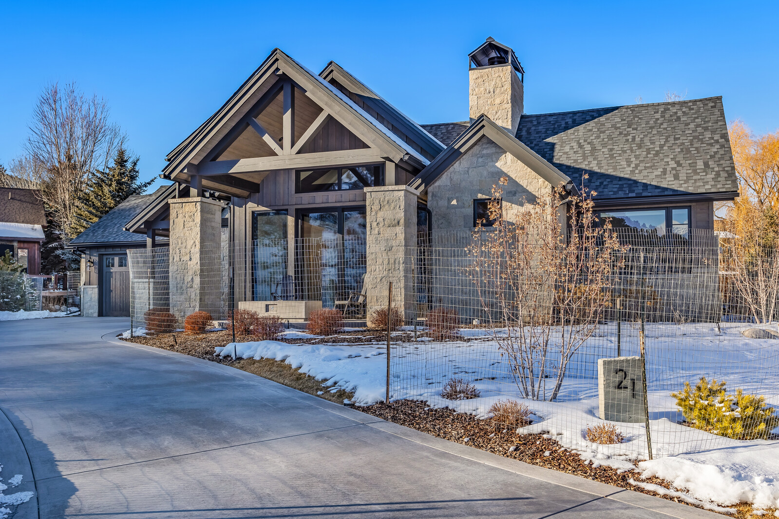 This stunning mountain-style residence features a sophisticated blend of natural stone and dark wood siding, highlighted by a prominent gabled roofline and a grand stone chimney. A concrete driveway leads up to the home, which is framed by a wire fence and light snow cover, creating a crisp, high-end aesthetic. The architectural design emphasizes a rustic yet modern luxury, perfectly suited for a scenic, cold-climate setting.