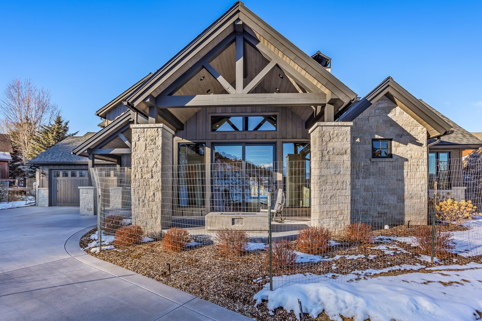 This stunning mountain-style home features a striking architectural design with a prominent gabled roof, stone masonry, and dark wood accents. The rear exterior showcases a covered patio area with a fire pit, large glass sliding doors, and a fenced-in yard set against a backdrop of snow-dusted mountains. The perspective is a wide-angle, eye-level shot that emphasizes the home's luxurious, rustic aesthetic and seamless integration with the natural landscape.