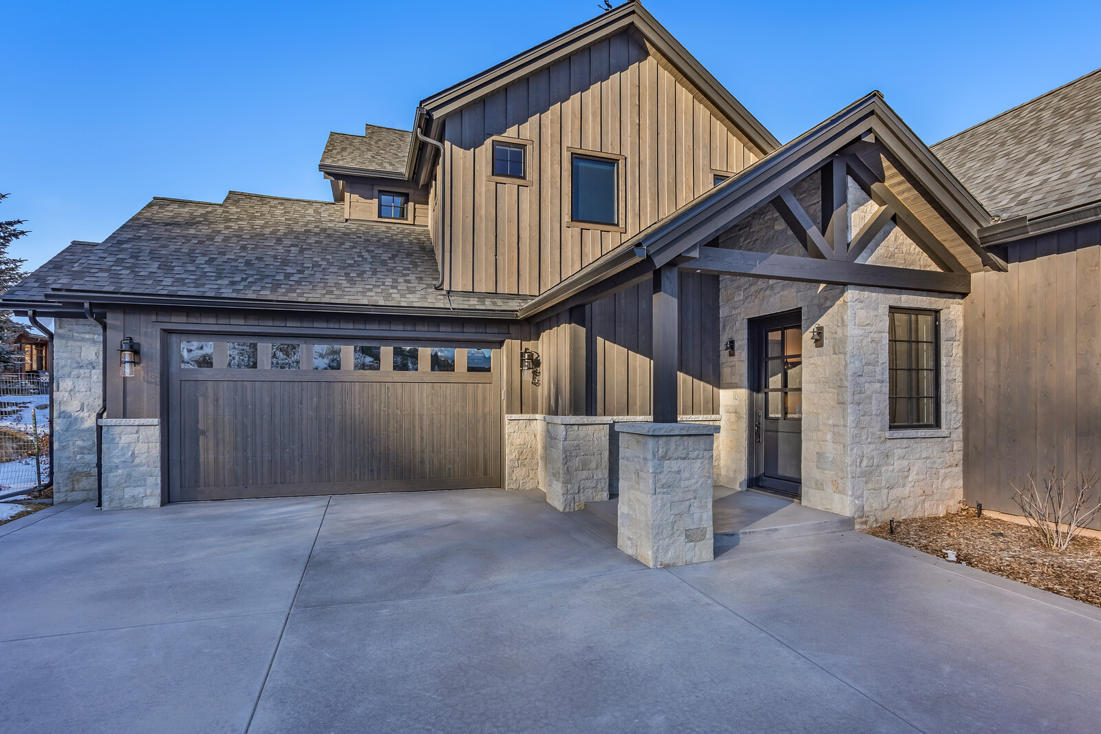 This striking modern farmhouse exterior features a harmonious blend of vertical wood siding and light-toned stone masonry. The home includes a spacious attached garage and a welcoming covered entryway with timber-framed accents, all set against a clear blue sky. The clean lines and high-contrast materials create a sophisticated, high-end curb appeal.