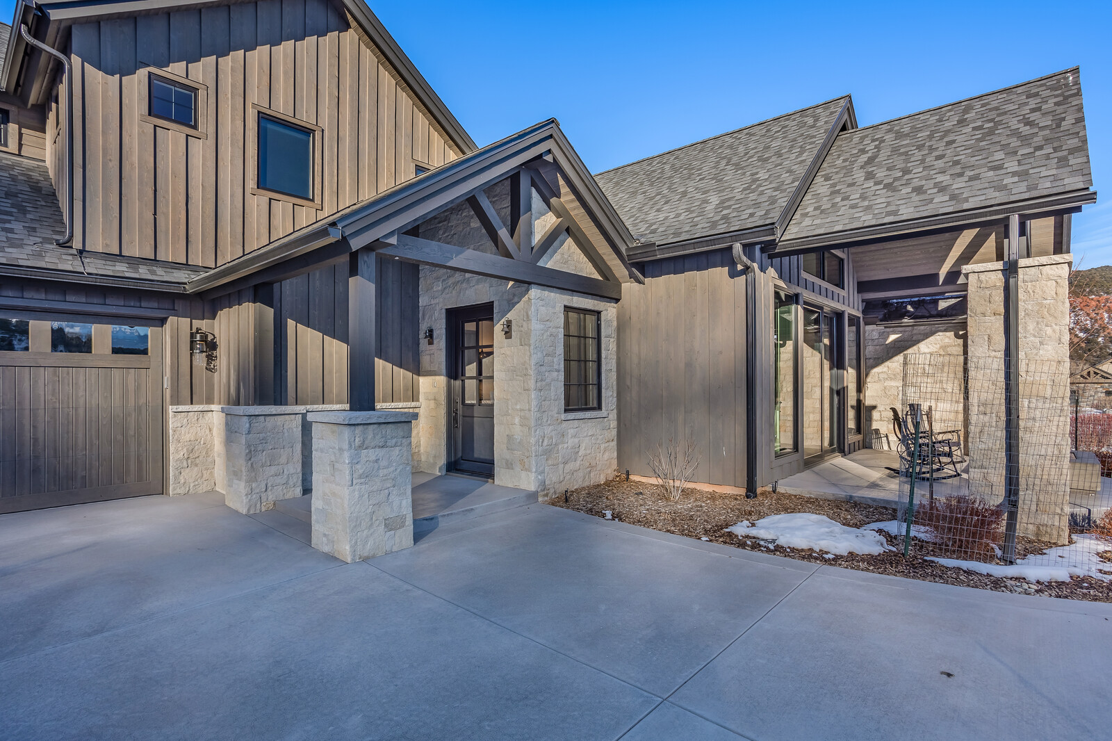 This striking modern farmhouse exterior features a blend of dark vertical board-and-batten siding and light-toned stone masonry. The entryway is highlighted by a timber-framed gable roof structure, leading to a glass-paneled front door, while an adjacent covered patio area provides a cozy outdoor seating space. The perspective is a wide-angle shot from the driveway, capturing the clean lines and architectural contrast of the home's facade under a clear blue sky.