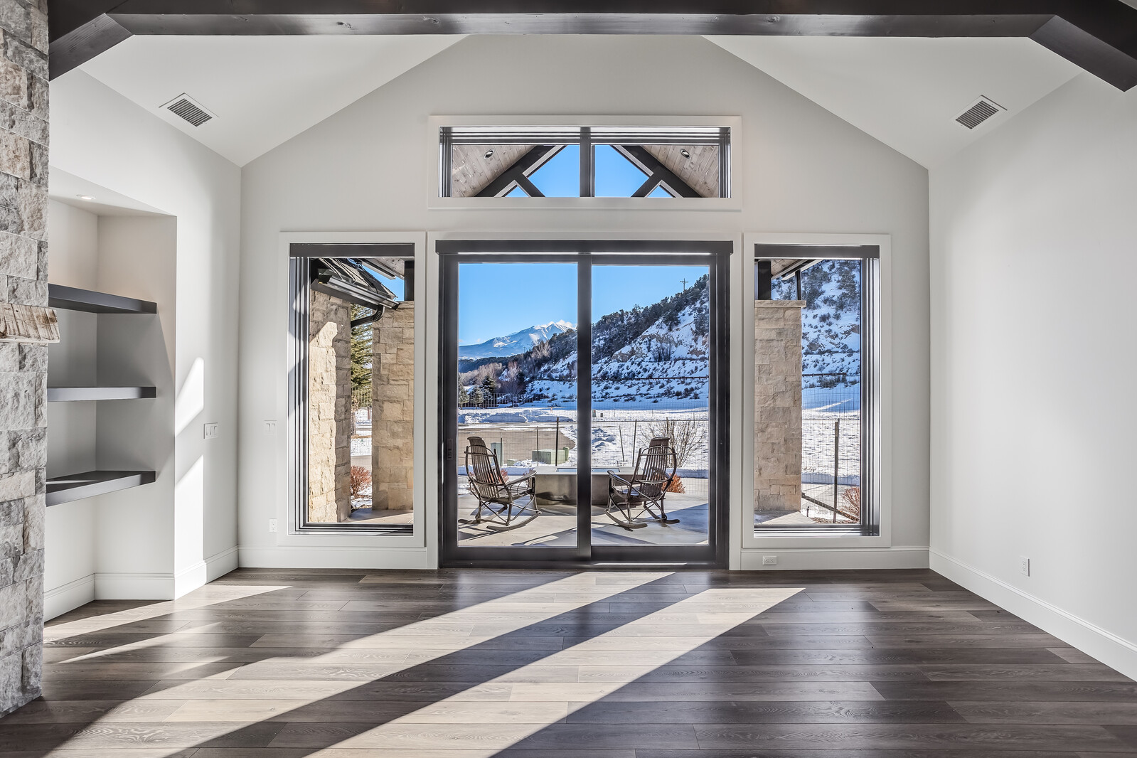 This bright and airy living room features a vaulted ceiling with dark wood beams and expansive floor-to-ceiling windows that frame a stunning mountain view. The space is anchored by dark wood flooring and a stone-accented wall with built-in shelving, creating a sophisticated, rustic-modern aesthetic. The sliding glass doors open onto a patio, seamlessly blending the indoor living area with the snowy outdoor landscape.