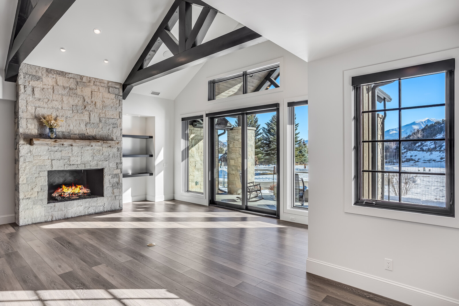 This spacious living room features a striking floor-to-ceiling stone fireplace with a rustic wooden mantel and a vaulted ceiling accented by dark timber beams. Large sliding glass doors and windows flood the room with natural light, offering expansive views of a snowy mountain landscape. The open-concept design, combined with warm wood flooring and clean white walls, creates a sophisticated and inviting mountain-modern atmosphere.