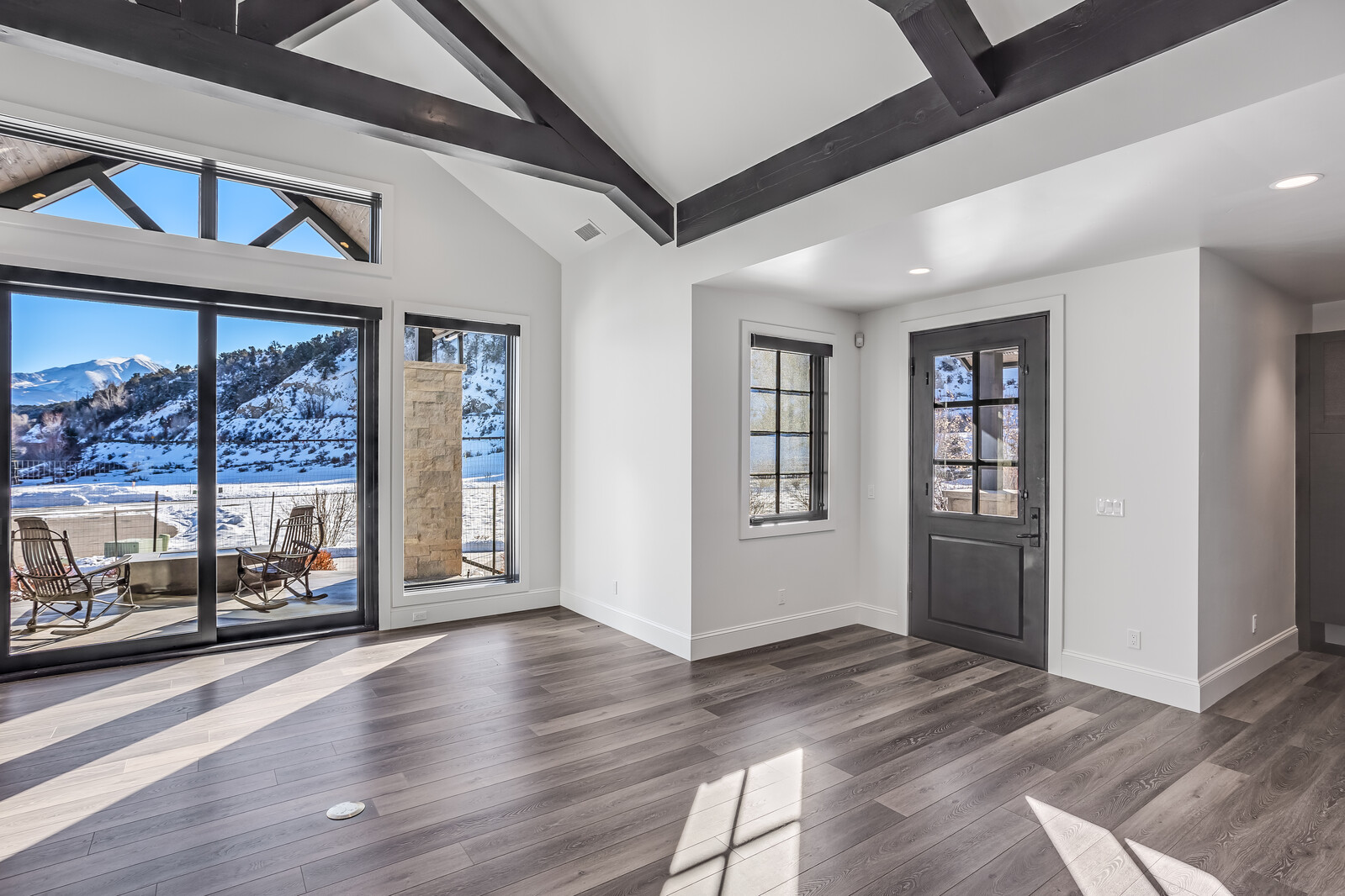 This bright and airy living room features high vaulted ceilings with exposed dark wood beams, creating a sophisticated mountain-modern aesthetic. Large sliding glass doors provide a seamless transition to an outdoor patio, framing a stunning, snow-capped mountain view. The space is finished with sleek, wood-look flooring and neutral white walls, offering a clean and inviting atmosphere.