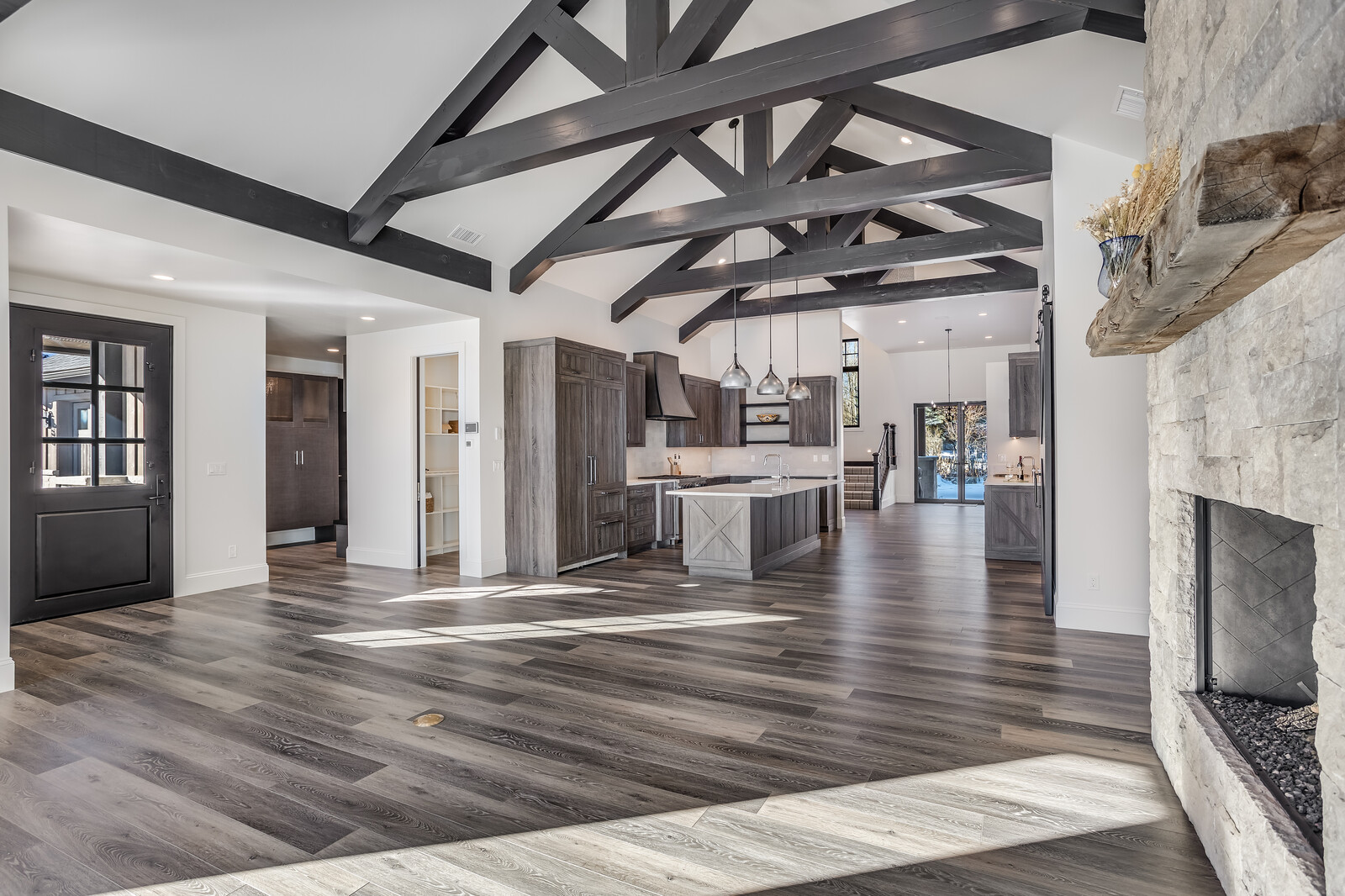 This expansive open-concept living area features a modern farmhouse aesthetic with dramatic dark wood vaulted ceiling trusses and wide-plank hardwood flooring. The kitchen is anchored by a large central island with a rustic X-pattern detail, complemented by dark wood cabinetry and sleek pendant lighting. A massive stone fireplace on the right adds texture and warmth, while the bright, airy layout flows seamlessly toward the rear glass doors.