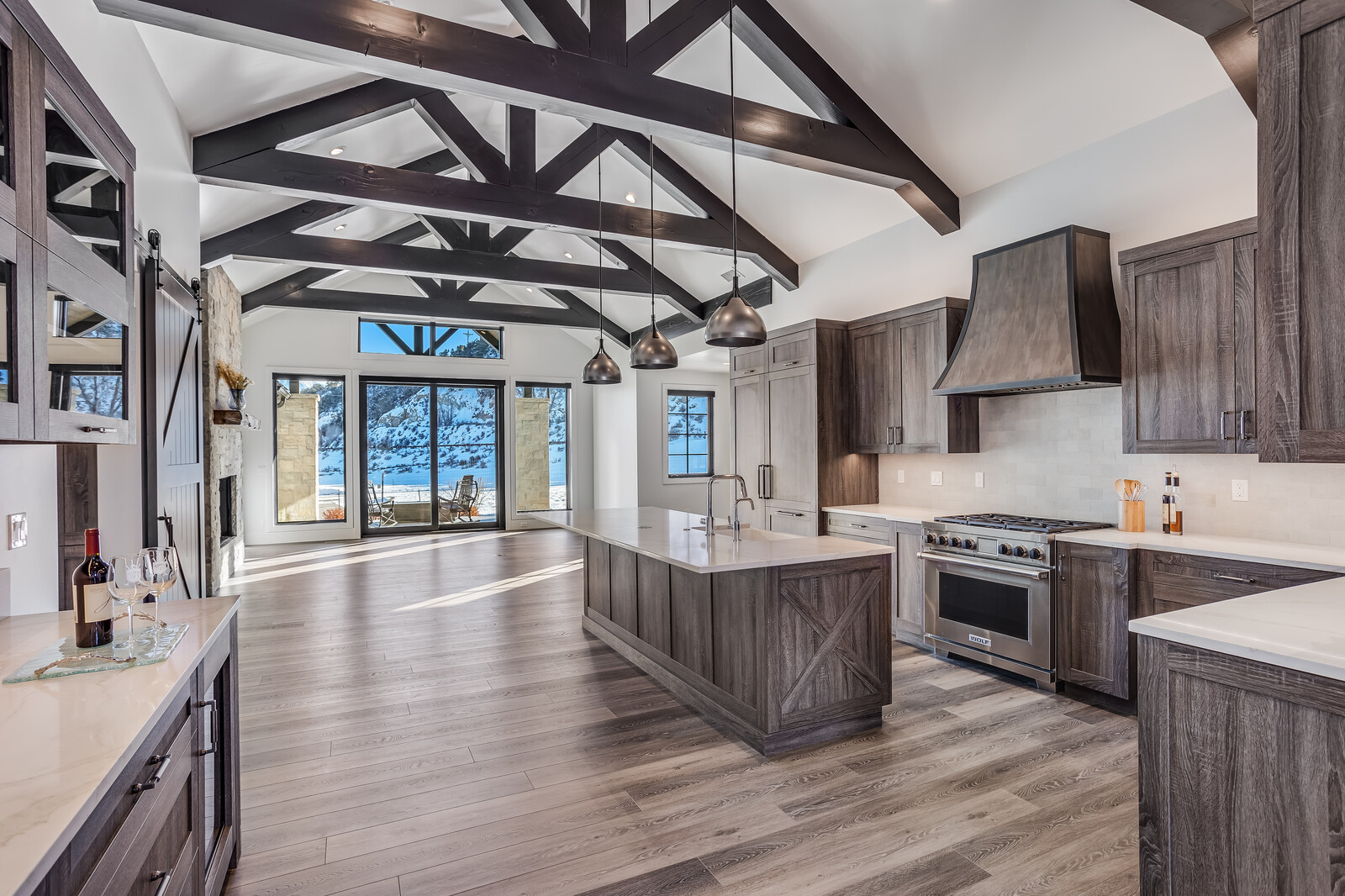 This spacious, high-end kitchen features a rustic-modern aesthetic with dark wood cabinetry, exposed timber ceiling beams, and a large central island with a quartz countertop. The open-concept design flows seamlessly toward a wall of glass doors that reveal a snowy outdoor landscape, creating a striking contrast between the warm interior and the cold exterior. Three industrial-style pendant lights hang above the island, highlighting the professional-grade stainless steel appliances and the sophisticated, clean lines of the space.