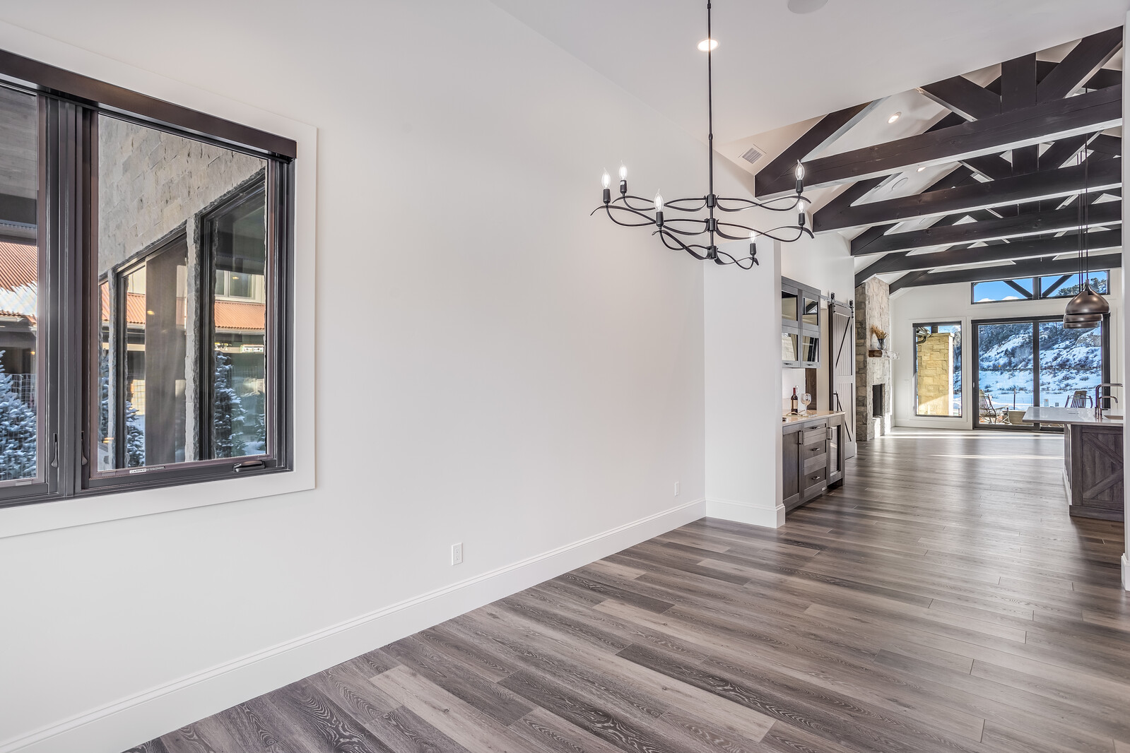 This spacious, open-concept dining area features elegant dark wood ceiling beams and high-quality grey-toned wood flooring. A sophisticated black chandelier serves as a focal point, while the room flows seamlessly into a kitchen area with a wet bar and a view of a snowy landscape through large windows. The bright, neutral walls and clean lines create a modern, airy atmosphere perfect for entertaining.