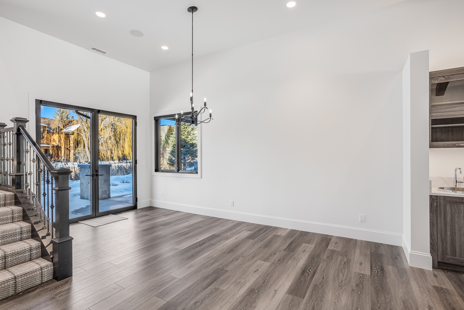 This bright and airy dining area features high ceilings, modern wood-look flooring, and a sleek black chandelier that serves as a focal point. A set of glass doors provides a seamless transition to the outdoor space, while a staircase with a dark wood railing adds architectural interest to the left. The room is clean and minimalist, offering a versatile space that connects to a small wet bar area on the right.