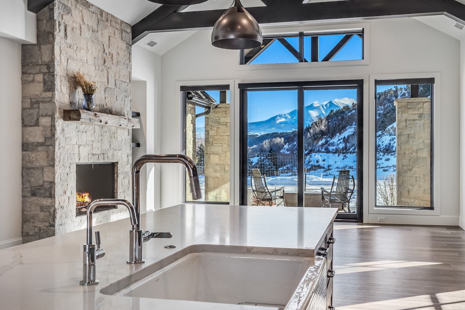 This bright, modern kitchen features a large white quartz island with a farmhouse sink and polished chrome faucet, serving as the focal point of the room. A stunning stone-clad fireplace adds warmth to the space, while expansive floor-to-ceiling windows frame a breathtaking, panoramic view of snow-capped mountains. The open-concept design, highlighted by dark wood beams and sleek pendant lighting, creates a luxurious and inviting atmosphere perfect for mountain living.