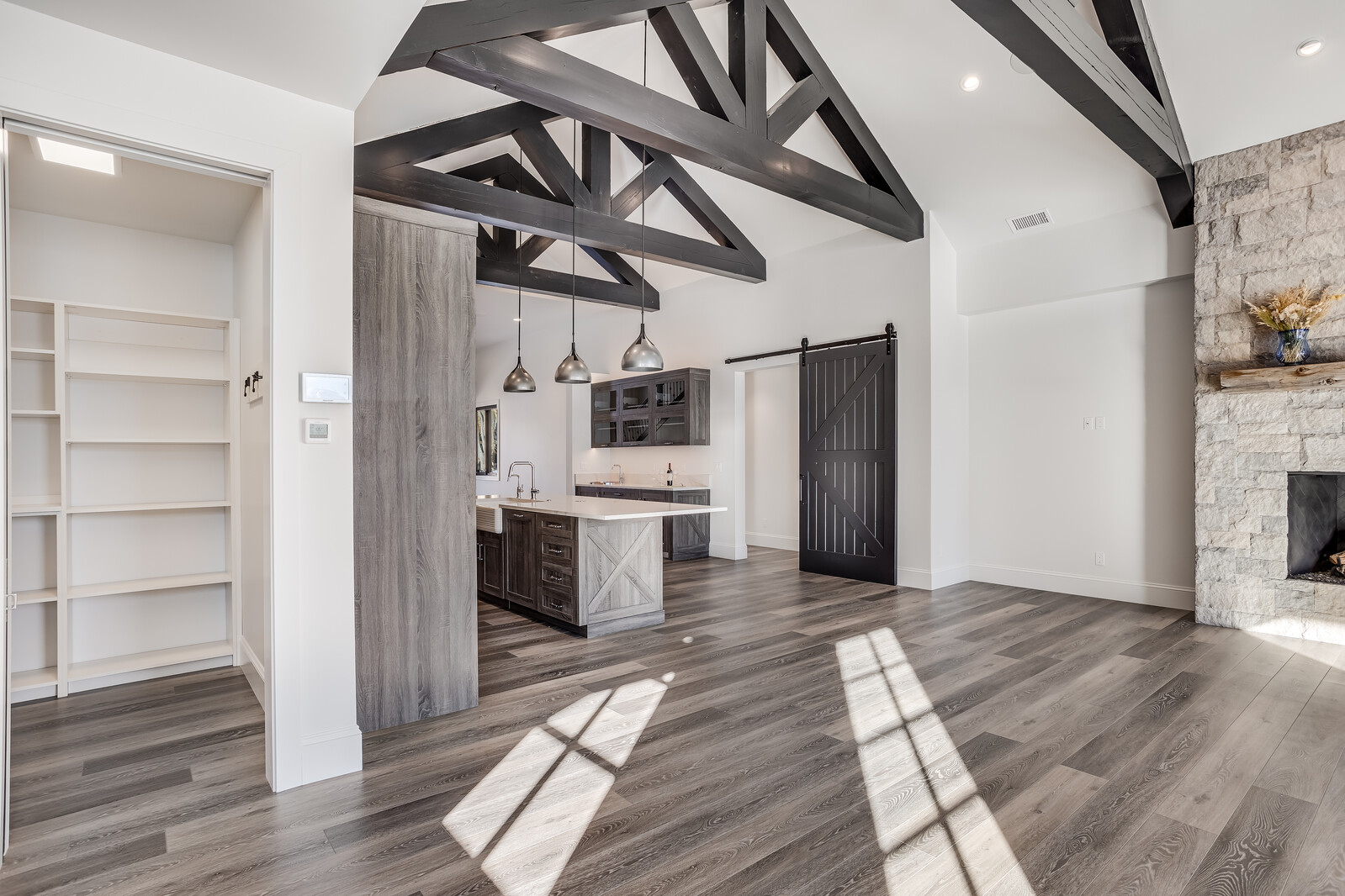 This spacious open-concept kitchen and living area features a striking vaulted ceiling with dark exposed wooden trusses and modern pendant lighting. The space showcases a blend of rustic and contemporary design, highlighted by a stone fireplace, a large kitchen island with a farmhouse sink, and a sliding barn door. The wide-angle perspective emphasizes the seamless flow between the kitchen, the adjacent pantry, and the expansive living area, all tied together by warm wood-look flooring.