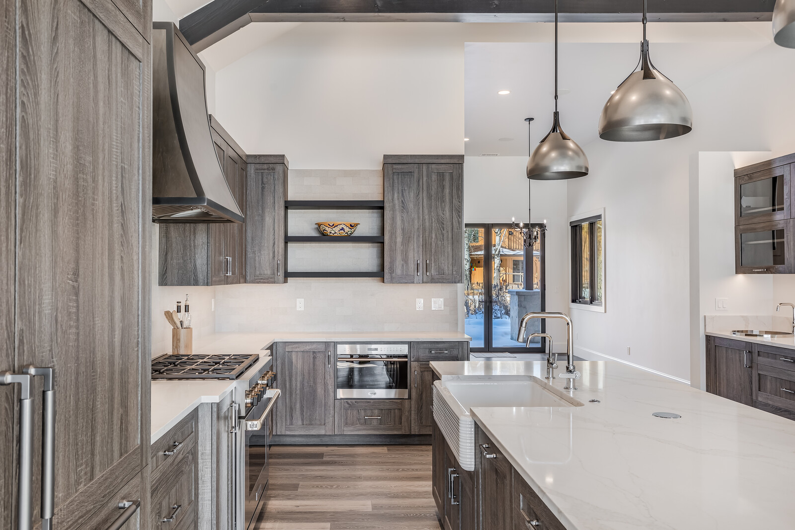 This modern, high-end kitchen features sleek, dark wood-grain cabinetry paired with pristine white quartz countertops and a farmhouse-style sink. The space is anchored by a large central island with pendant lighting and is equipped with professional-grade stainless steel appliances, including a gas range and built-in oven. The open-concept design flows seamlessly toward a bright, snow-covered outdoor view, creating a sophisticated and inviting atmosphere.