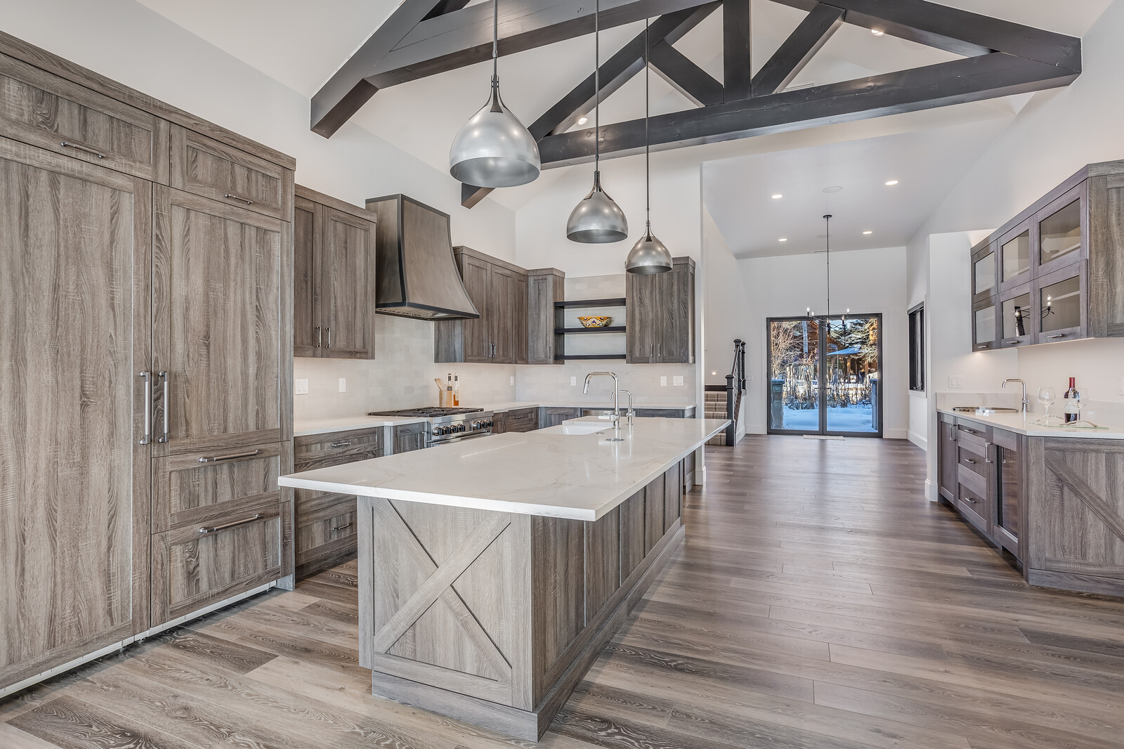 This spacious, modern farmhouse-style kitchen features rustic wood-toned cabinetry, a large central island with a white quartz countertop, and striking dark exposed ceiling beams. The open-concept design flows seamlessly into a bright dining area with glass doors that reveal a snowy outdoor view, creating a sophisticated yet cozy atmosphere. Three metallic pendant lights hang above the island, providing a focal point that complements the high-end stainless steel appliances and clean, neutral aesthetic.
