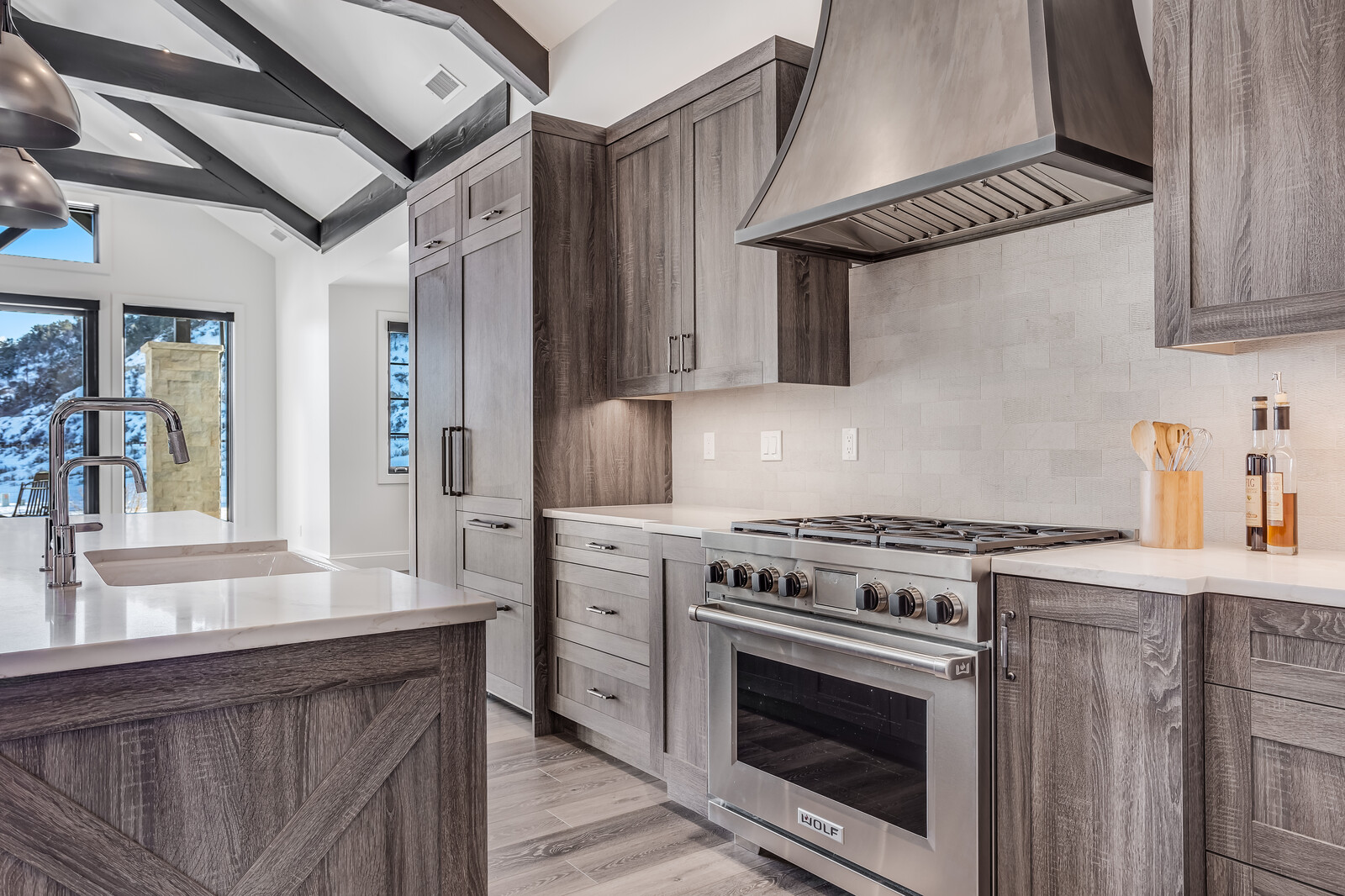 This sophisticated kitchen features rustic-modern aesthetics with dark wood-grained cabinetry, a professional-grade Wolf range, and a sleek stainless steel hood. The space is anchored by a large kitchen island with a white marble countertop and a farmhouse-style sink, all set beneath a dramatic vaulted ceiling with exposed dark beams. Large windows provide a bright, scenic view of a snowy landscape, creating a luxurious and cozy mountain-home atmosphere.