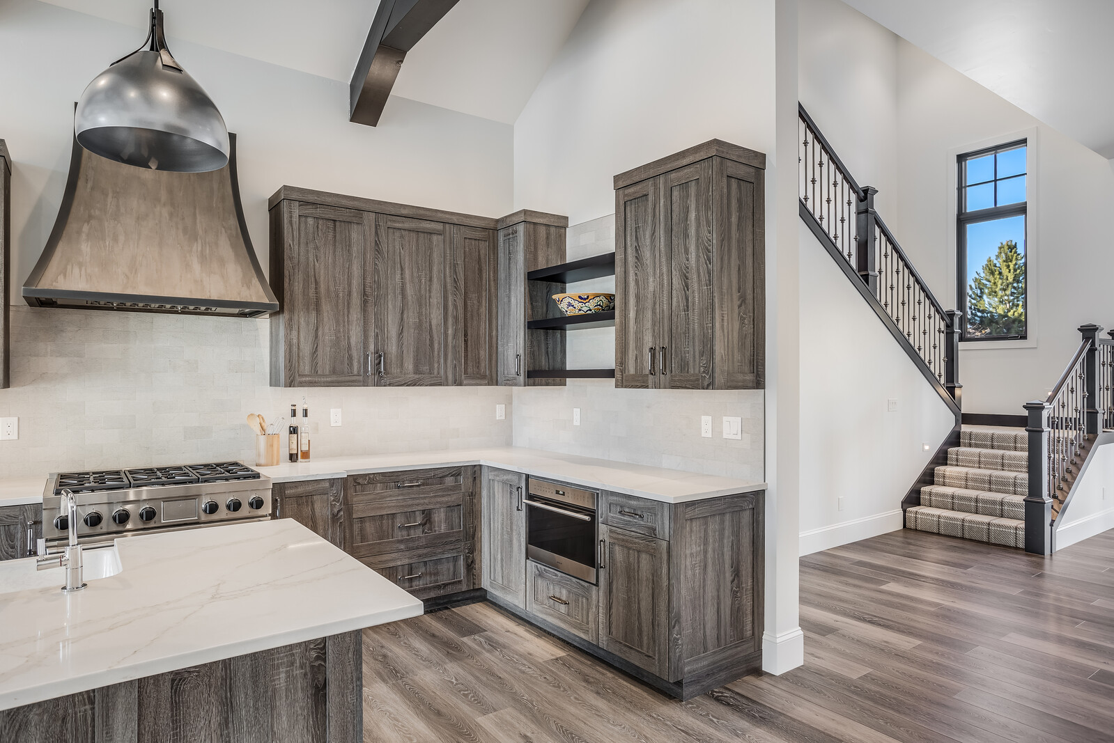 This modern kitchen features rustic wood-grain cabinetry paired with sleek white quartz countertops and a matching backsplash. A prominent metal range hood hangs above the gas stove, while an open staircase with a black iron railing adds architectural interest to the adjacent hallway. The space is bright and airy, showcasing a sophisticated blend of contemporary design and warm, textured finishes.
