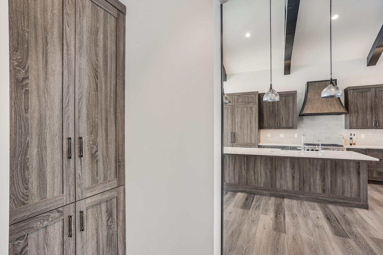 This image captures a view from a hallway looking into a modern, open-concept kitchen featuring dark wood-grain cabinetry and a large central island with a white countertop. The space is accented by sleek pendant lighting, a custom range hood, and exposed ceiling beams, creating a sophisticated and cohesive aesthetic. The perspective emphasizes the transition between the storage area in the foreground and the expansive, well-lit kitchen beyond.