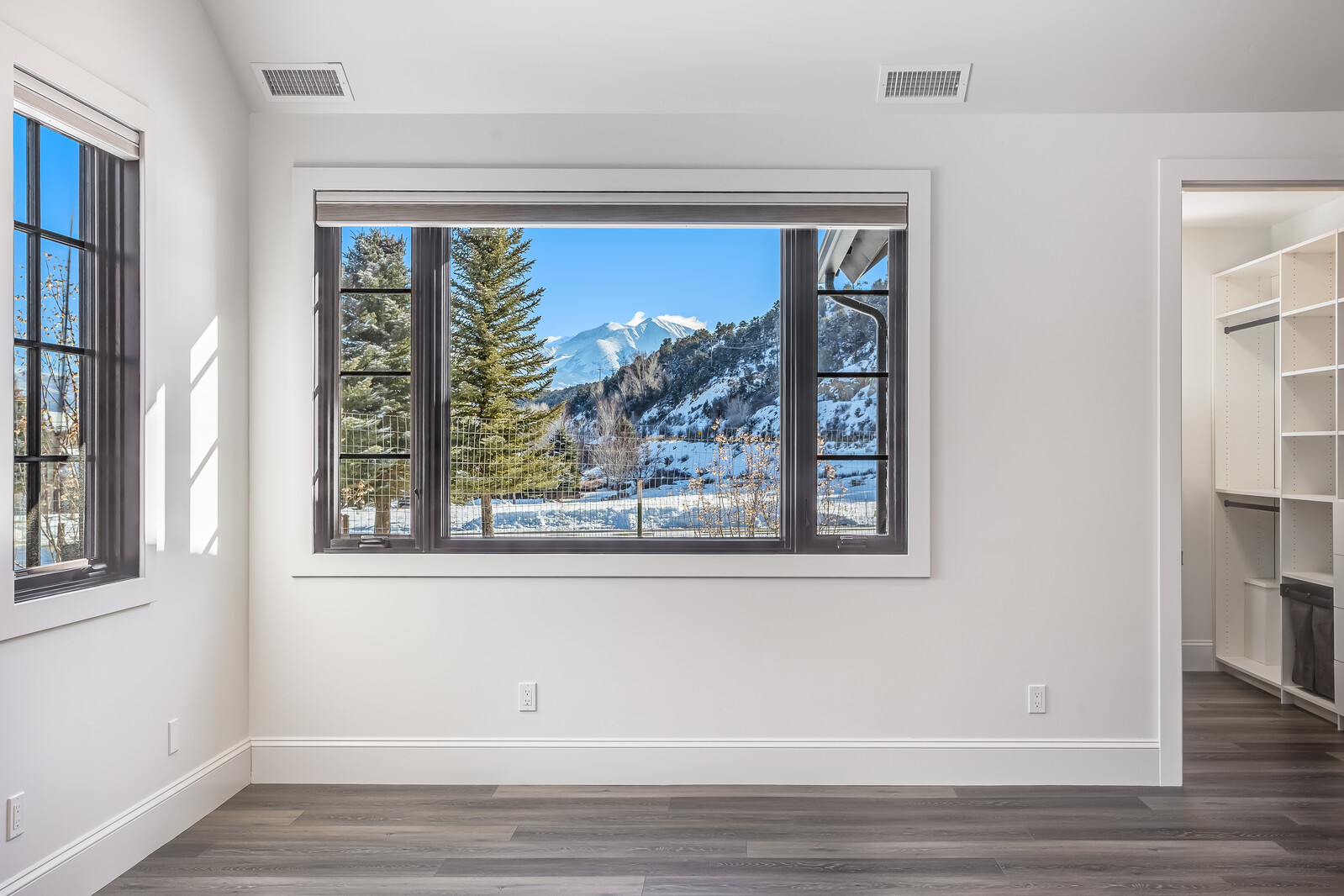 This bright and minimalist bedroom features crisp white walls and modern dark wood-look flooring, creating a clean and contemporary aesthetic. The room is anchored by a large, dark-framed window that serves as a focal point, showcasing a stunning, unobstructed view of snow-capped mountains and evergreen trees. To the right, an open doorway leads into a spacious, well-organized walk-in closet with built-in shelving, emphasizing the home's functional design.