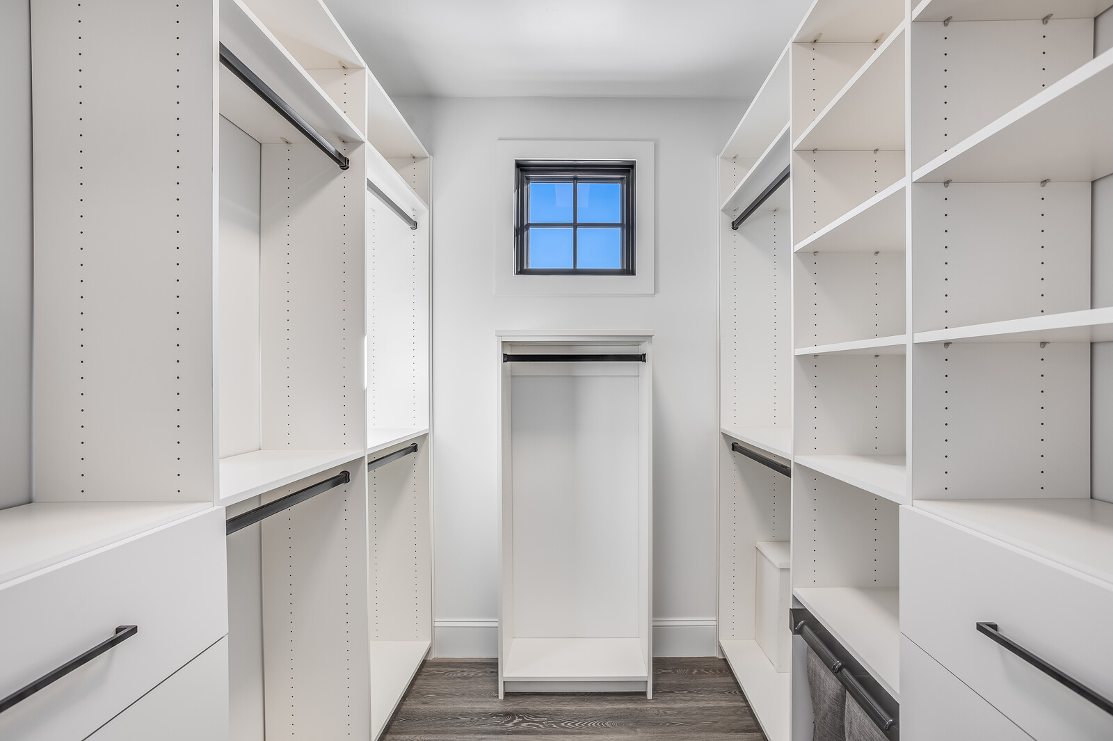 This walk-in closet features a clean, modern design with extensive white custom shelving and hanging rods on both sides. A small, centered window allows natural light to brighten the space, which is finished with dark wood-look flooring. The symmetrical layout and minimalist aesthetic create an organized and spacious feel.