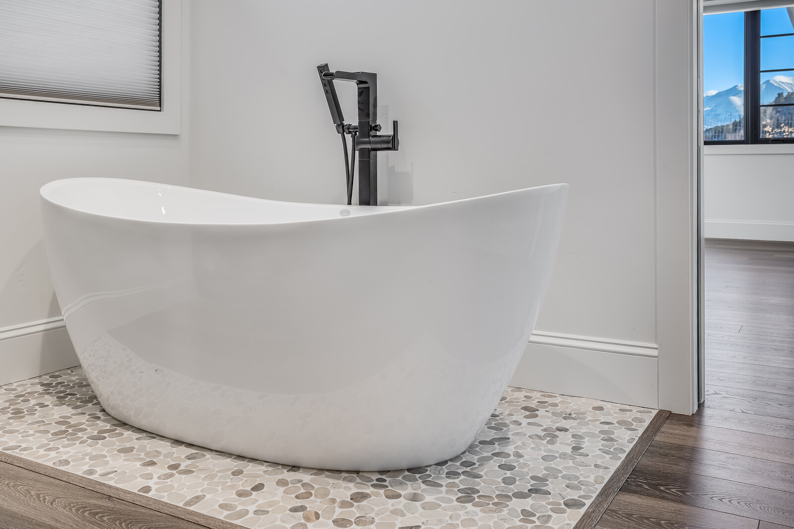 This elegant bathroom features a sleek, white freestanding soaking tub as the centerpiece, complemented by a modern matte black floor-mounted faucet. The tub sits on a textured pebble-stone floor inlay that contrasts beautifully with the surrounding dark wood-look flooring. A window with a view of snow-capped mountains adds a serene, high-end touch to the minimalist, contemporary design.