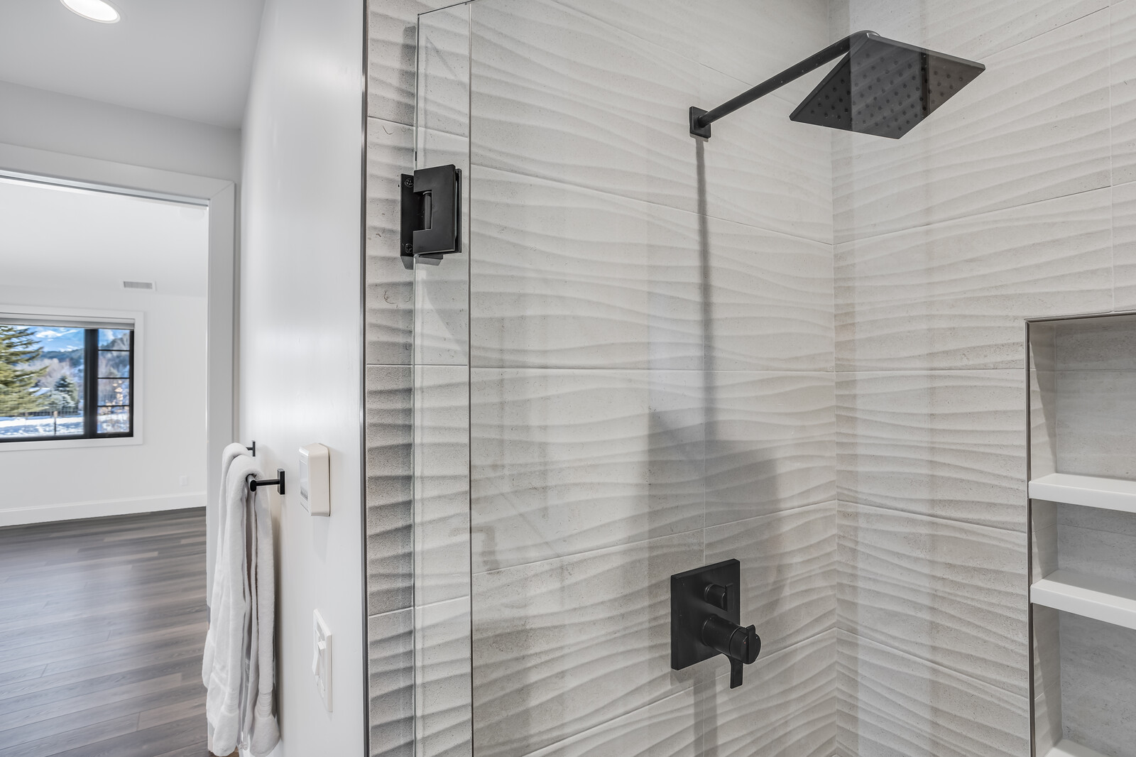 This close-up shot highlights a modern, high-end shower enclosure featuring textured, wave-patterned wall tiles and sleek matte black fixtures. A glass door with a heavy-duty black hinge provides a clean, contemporary aesthetic, while a built-in recessed shelf offers functional storage. The perspective captures the transition from the tiled shower area to a bright, open room beyond, emphasizing the home's sophisticated design.
