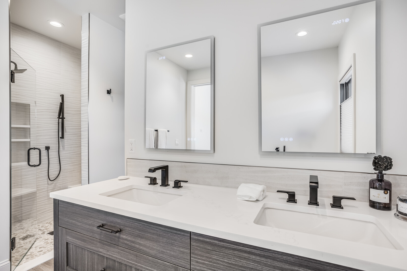 This modern primary bathroom features a sleek double vanity with a white quartz countertop, dark wood-grain cabinetry, and matte black fixtures. Two rectangular LED-lit mirrors hang above the sinks, while a glass-enclosed walk-in shower with pebble flooring and a rainfall showerhead is visible to the left. The space is bright and contemporary, emphasizing clean lines and a sophisticated, high-end aesthetic.