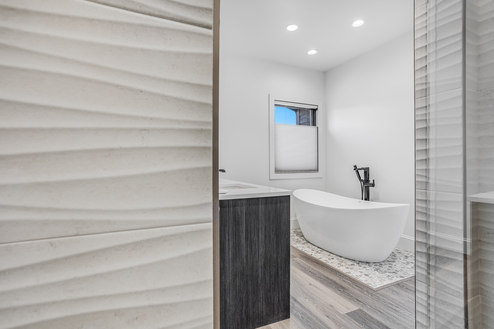 This modern primary bathroom features a sleek, freestanding soaking tub set against a backdrop of a pebble-tiled floor and a window with a pleated shade. To the left, a dark wood vanity with a white countertop adds a sophisticated contrast, while the foreground is framed by a textured, wavy wall panel. The perspective is captured from a doorway, creating a sense of depth and inviting the viewer into this serene, spa-like retreat.