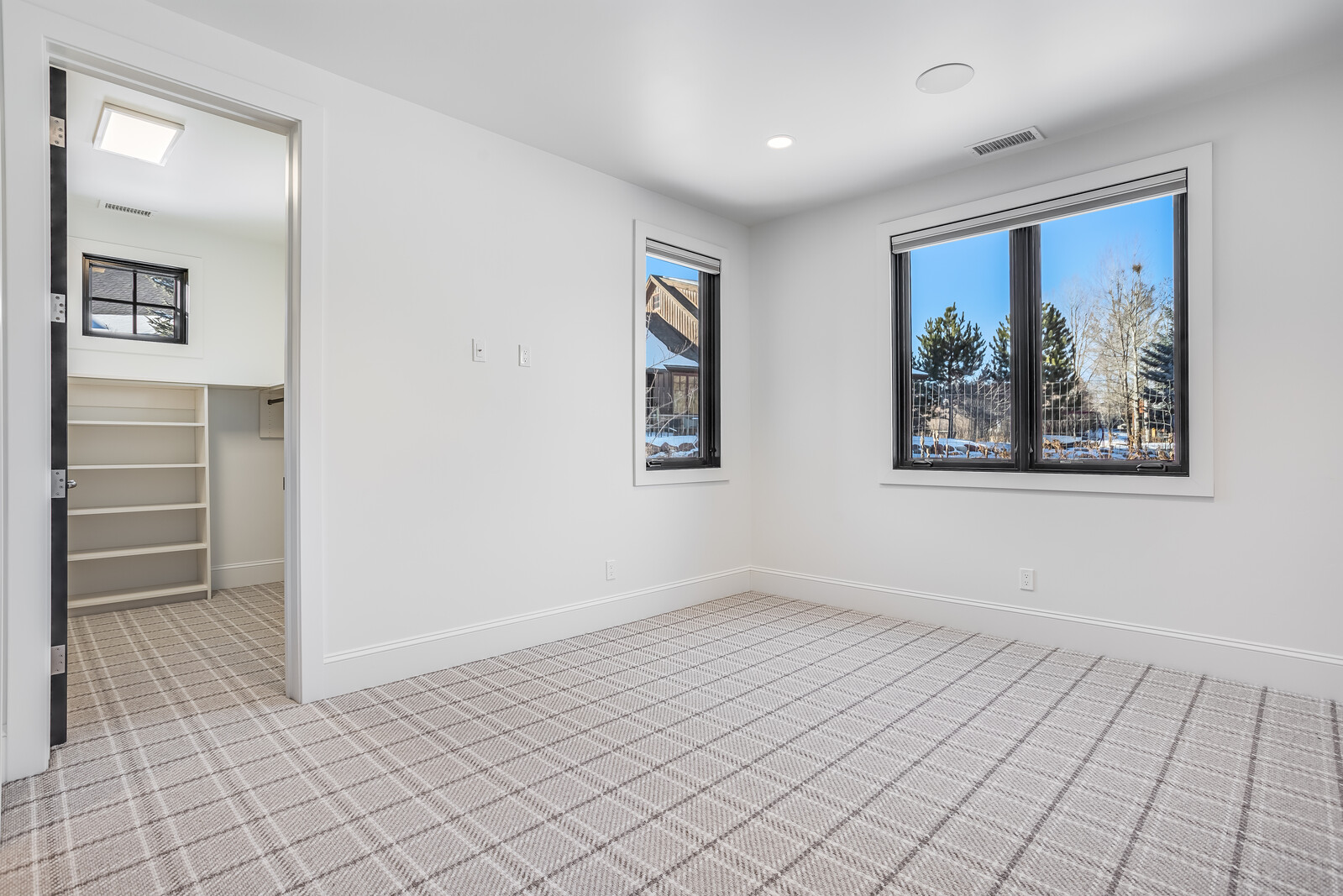 This bright and minimalist bedroom features crisp white walls, a neutral plaid-patterned carpet, and large windows that provide ample natural light and scenic outdoor views. An open doorway leads into a walk-in closet equipped with built-in shelving, enhancing the room's functional appeal. The clean, modern aesthetic creates a versatile and inviting space suitable for a guest room or home office.