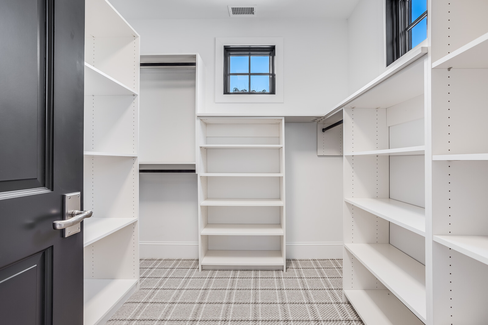This walk-in closet features a clean, modern aesthetic with crisp white custom shelving and hanging rods arranged in a functional U-shaped layout. The space is grounded by a stylish grey and white plaid patterned carpet, while a central window allows natural light to brighten the room. The dark grey door provides a sophisticated contrast to the bright, organized storage area.