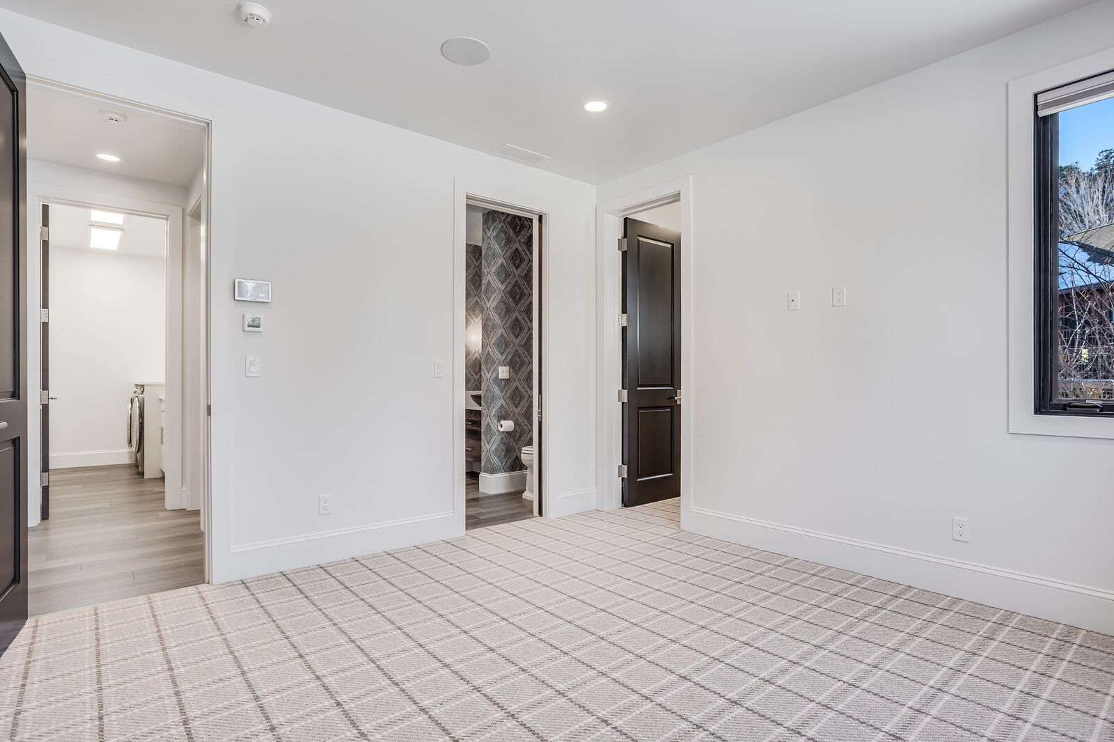 This bright and neutral bedroom features a clean, modern aesthetic with light-colored plaid carpeting and crisp white walls. The room includes two doorways leading to adjacent spaces, one of which reveals a stylish bathroom with patterned wallpaper, and a large window that allows for natural light. The perspective is centered, showcasing the room's functional layout and potential for versatile use.