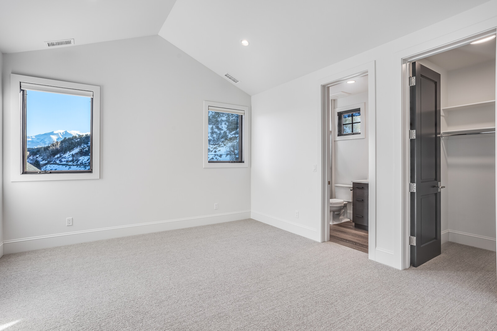 This bright and airy bedroom features vaulted ceilings, neutral carpeting, and crisp white walls, creating a clean and inviting atmosphere. Two windows provide ample natural light and offer scenic views of snow-capped mountains, while an open doorway leads to an en-suite bathroom and a walk-in closet. The minimalist design and high-quality finishes suggest a modern, well-maintained living space.