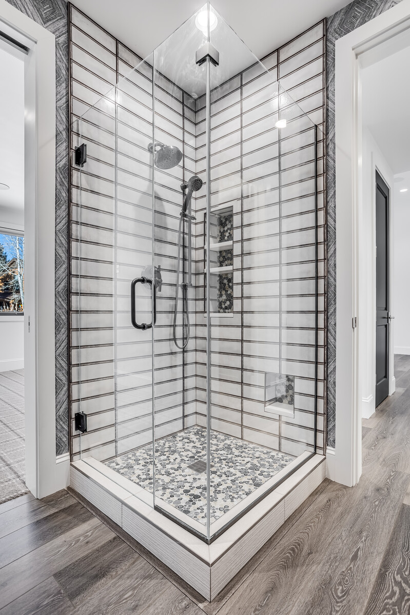 This modern, high-end walk-in shower features a striking combination of elongated white subway tiles with dark grout and a textured pebble-stone floor. The glass-enclosed space includes a built-in vertical niche for storage, a rainfall showerhead, and a handheld sprayer, all finished in sleek black hardware. The perspective is a clean, eye-level shot that highlights the sophisticated contrast between the geometric wall patterns and the organic floor texture.