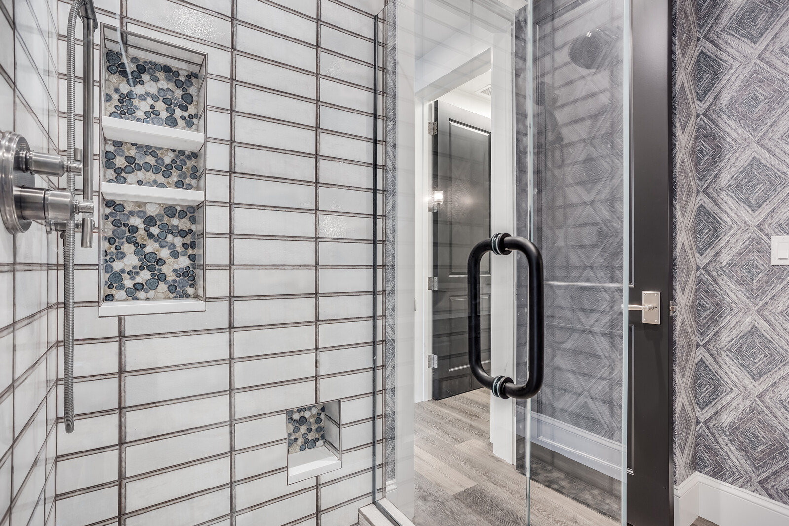 This modern bathroom features a sleek glass-enclosed shower with white subway tiles and decorative pebble-mosaic recessed shelving. The space is accented by a dark, patterned wallpaper and a contemporary black door handle, creating a sophisticated and high-end aesthetic. The perspective captures the intricate tile work and the transition into the adjacent room, highlighting the clean lines and premium finishes.