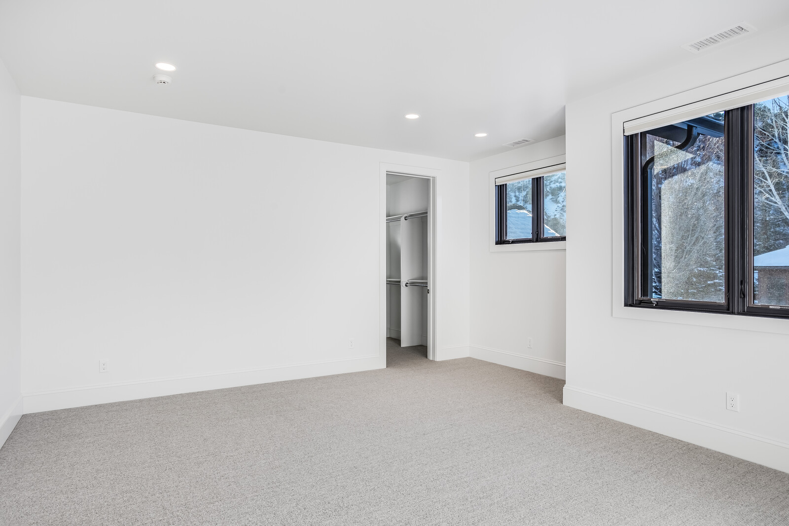 This bright and minimalist bedroom features crisp white walls, neutral carpeting, and a clean, modern aesthetic. The room includes a walk-in closet and large windows with dark frames that provide a view of the snowy exterior, creating a serene and inviting atmosphere. The perspective is a wide-angle shot from the corner of the room, emphasizing the spaciousness and natural light.