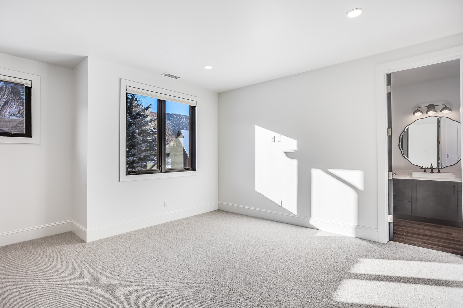 This bright and minimalist bedroom features neutral gray carpeting and crisp white walls, creating a clean and inviting atmosphere. A large window allows for natural light to flood the space, while an open doorway leads directly into a modern, well-appointed bathroom. The room's simple, uncluttered design offers a versatile canvas for potential residents.