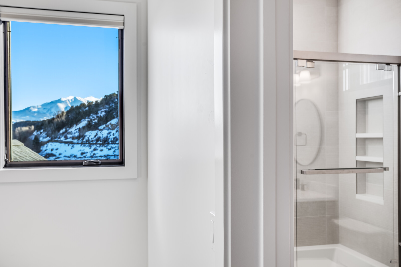 This interior shot captures a clean, modern bathroom transition, featuring a glass-enclosed shower with built-in shelving and a bright, white-walled room. To the left, a window provides a stunning, cinematic view of snow-capped mountains, creating a serene and luxurious atmosphere. The composition highlights the contrast between the minimalist interior design and the rugged natural beauty outside.