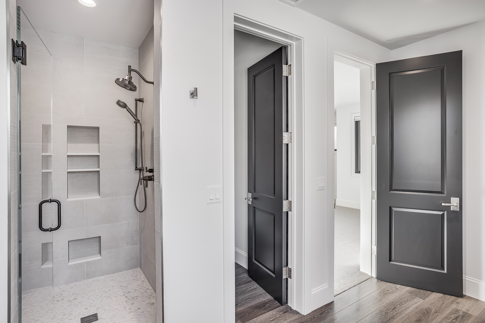 This modern bathroom features a sleek, glass-enclosed walk-in shower with light-toned tile, built-in wall niches, and a pebble-stone floor. The space transitions into a hallway with dark-toned wood flooring and two elegant, dark-painted doors that provide a striking contrast against the crisp white walls. The perspective captures a clean, contemporary design aesthetic that emphasizes high-end finishes and architectural detail.
