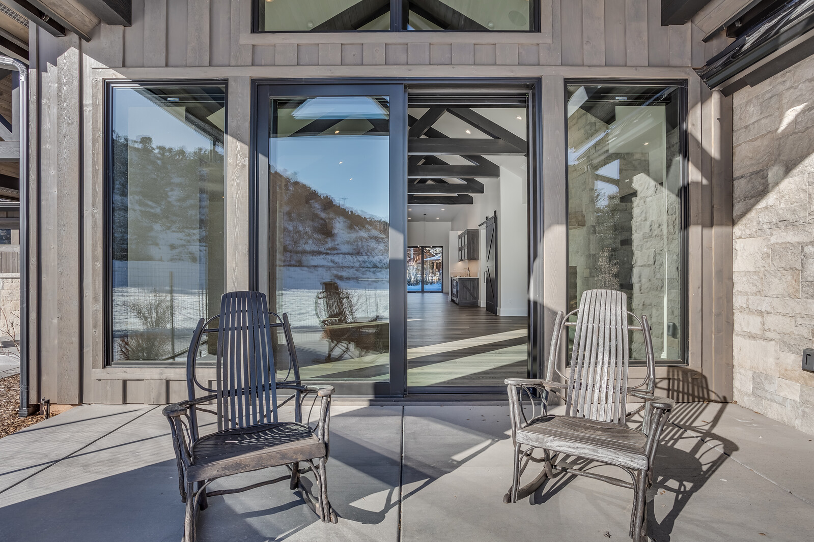 This image captures a serene patio space featuring two rustic wooden rocking chairs positioned on a smooth concrete surface. Large sliding glass doors provide a seamless transition to the home's interior, showcasing a vaulted ceiling with exposed dark beams and a view through to the back of the house. The setting offers a picturesque view of a snow-covered landscape, emphasizing a modern mountain aesthetic.