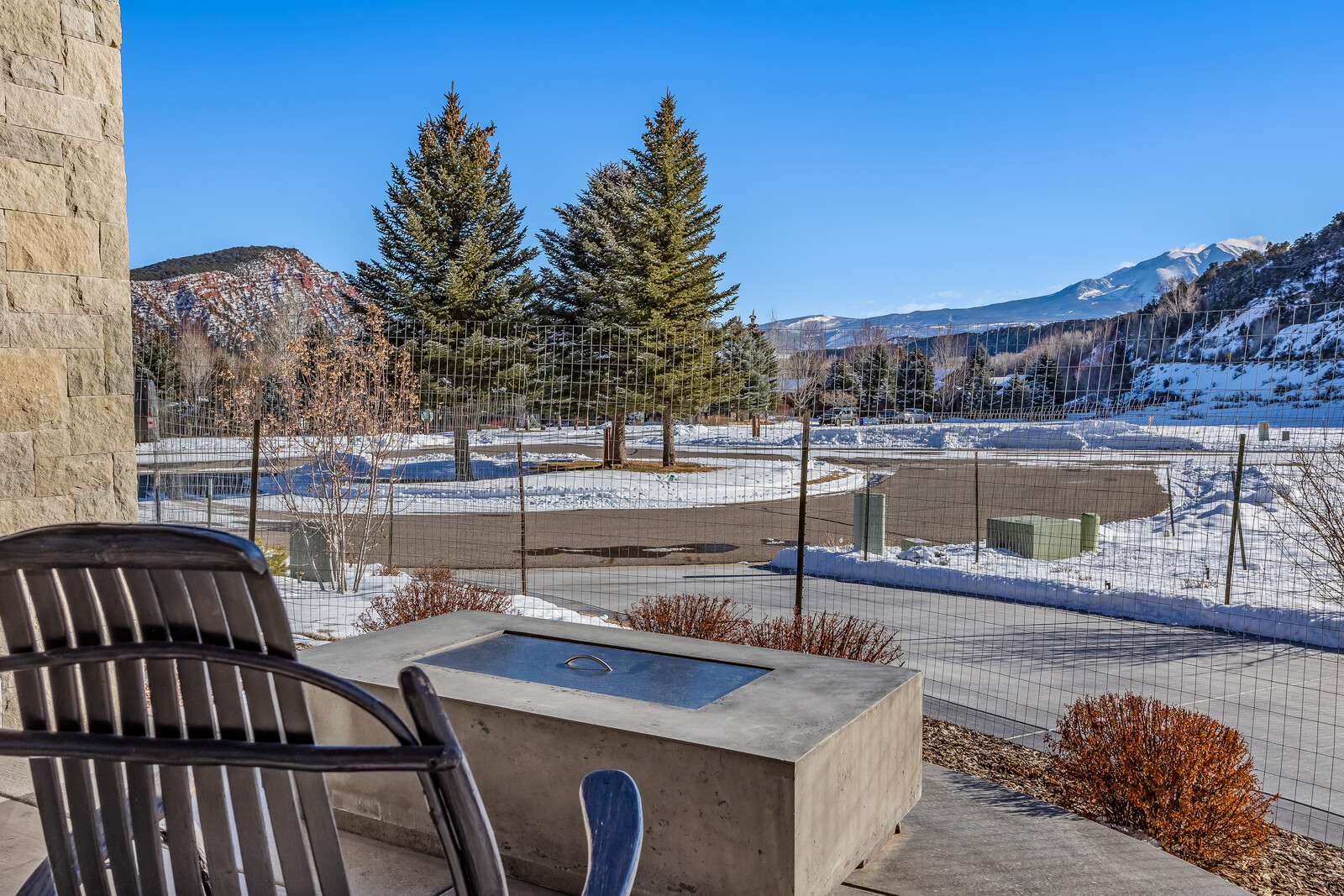 This image captures a serene outdoor patio space featuring a modern concrete fire pit and a comfortable wooden chair, perfect for enjoying the scenic mountain views. The perspective is from a slightly elevated vantage point, looking out through a wire fence toward a snow-dusted landscape with evergreen trees and distant peaks under a clear blue sky. The setting evokes a peaceful, high-altitude retreat atmosphere, highlighting the property's connection to its natural surroundings.