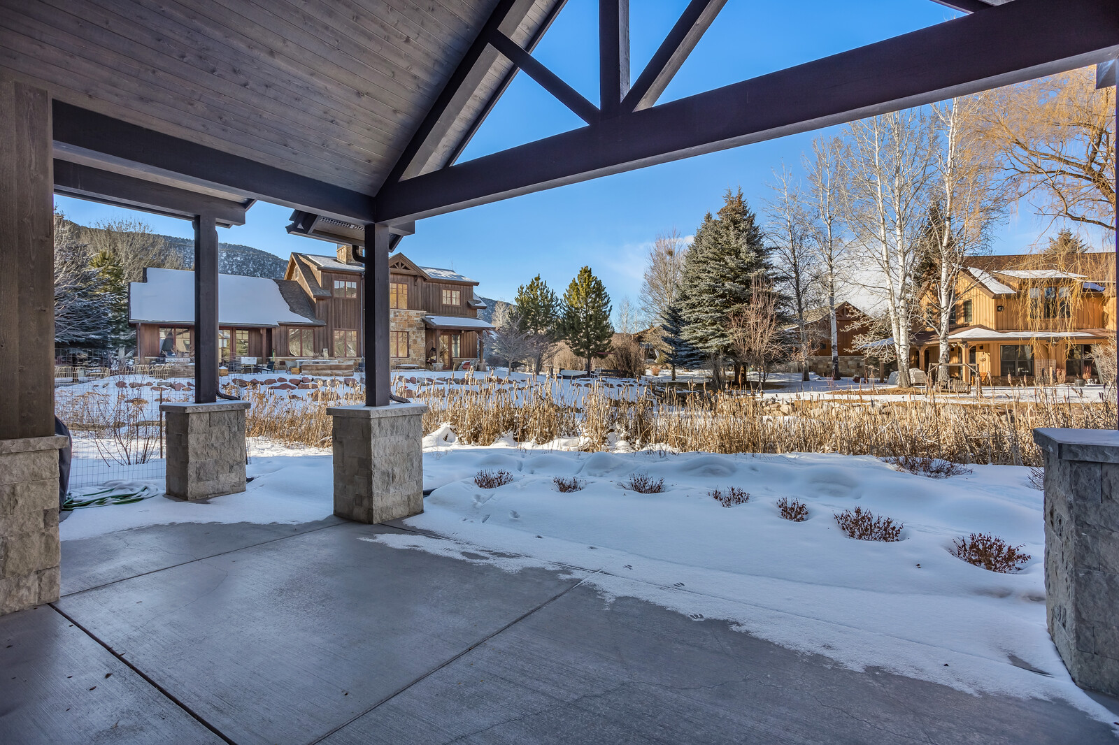 This image captures a serene, covered patio area looking out over a snow-covered landscape with a rustic, mountain-style home in the background. The structure features sturdy stone-based pillars and a vaulted timber-framed ceiling, creating a cozy and sheltered vantage point. The scene evokes a peaceful winter atmosphere, highlighting the property's connection to its natural, scenic surroundings.