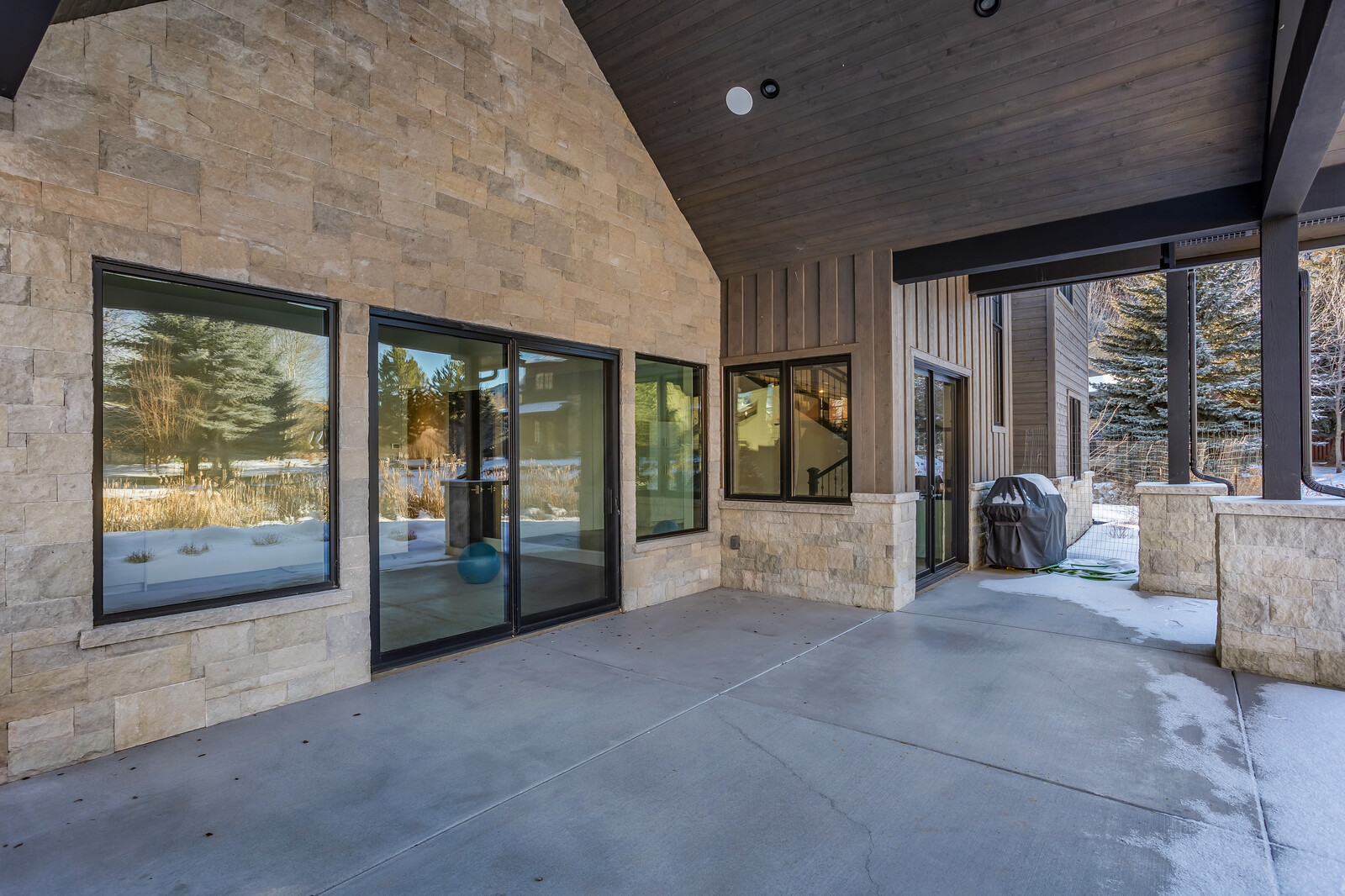 This image showcases a spacious, covered concrete patio featuring a blend of rustic stone masonry and modern dark wood siding. Large glass sliding doors provide a seamless transition between the indoor and outdoor living spaces, while the covered area offers a sheltered spot for outdoor dining or relaxation. The scene is set in a serene, snowy environment, highlighting the home's architectural appeal and year-round usability.