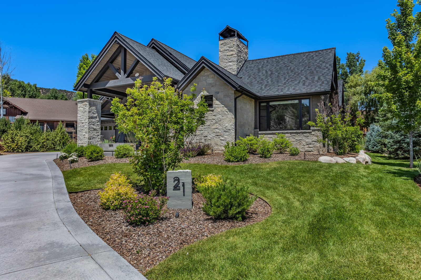 This stunning mountain-style home features a sophisticated blend of natural stone masonry and dark timber accents under a gabled roofline. A manicured lawn and lush landscaping frame the property, while a stone address marker labeled '21' sits prominently in the foreground. The bright blue sky and clear daylight emphasize the home's architectural elegance and inviting curb appeal.