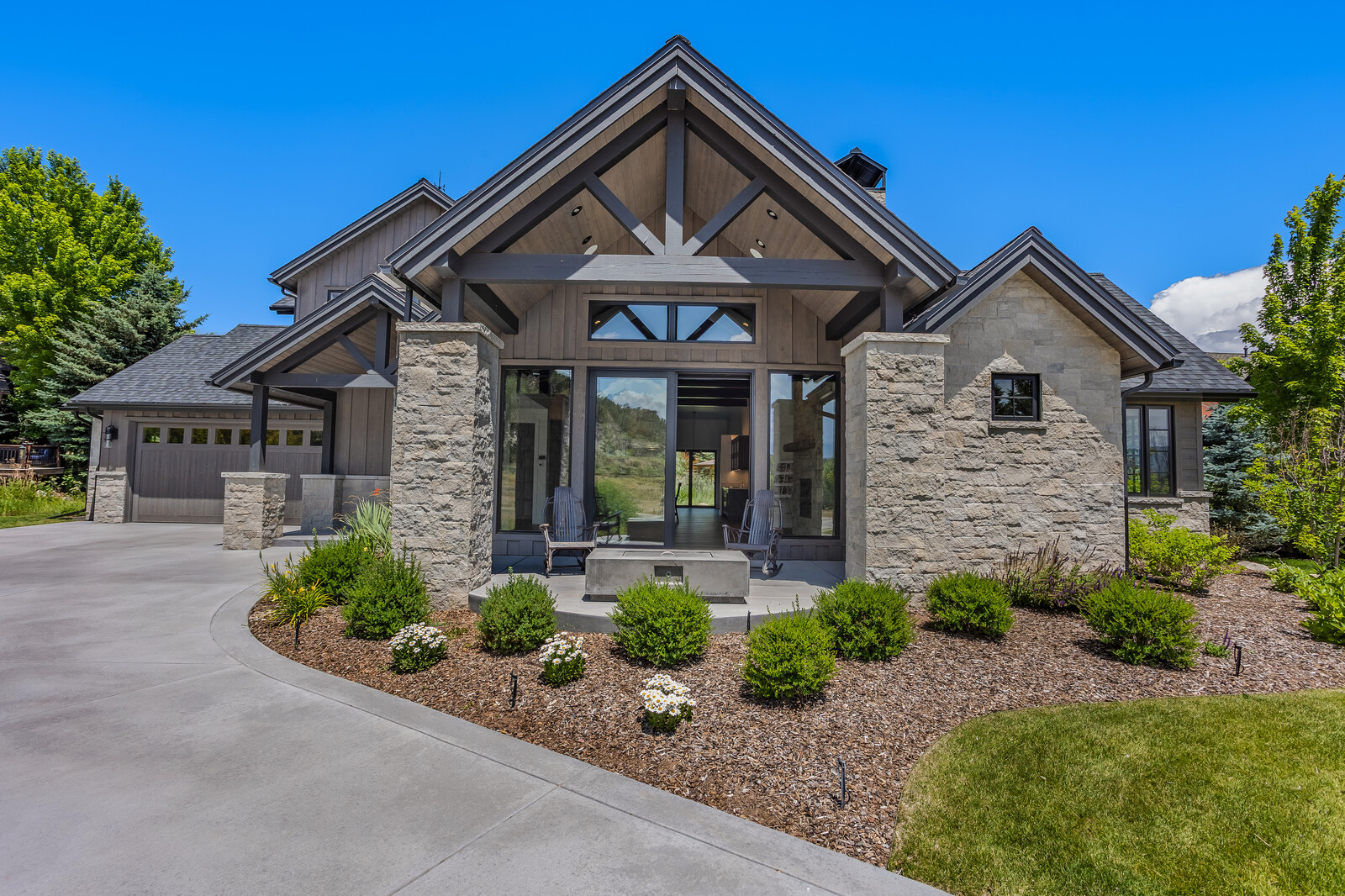 This stunning modern farmhouse features a striking combination of natural stone masonry and dark wood siding, accented by a prominent gabled roofline. The front entryway is framed by a covered porch with a stone fire pit, creating an inviting and sophisticated curb appeal. The meticulously landscaped garden beds and expansive concrete driveway enhance the home's clean, high-end aesthetic.
